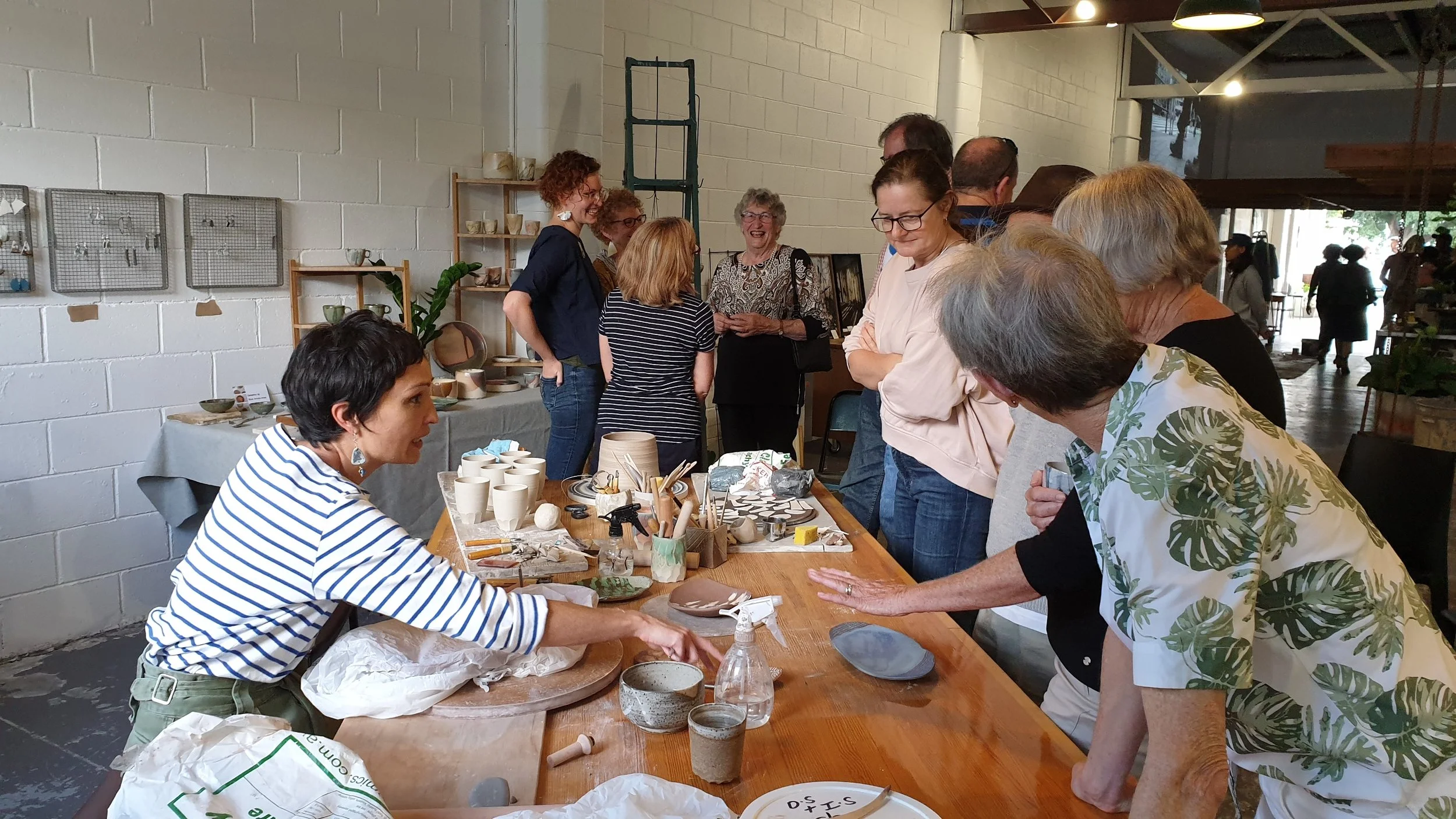 In the foreground a women is in conversation about making ceramics across a table, which is full of potters tools and some cups in their initial stages.  in the background are four women talking and laughing.