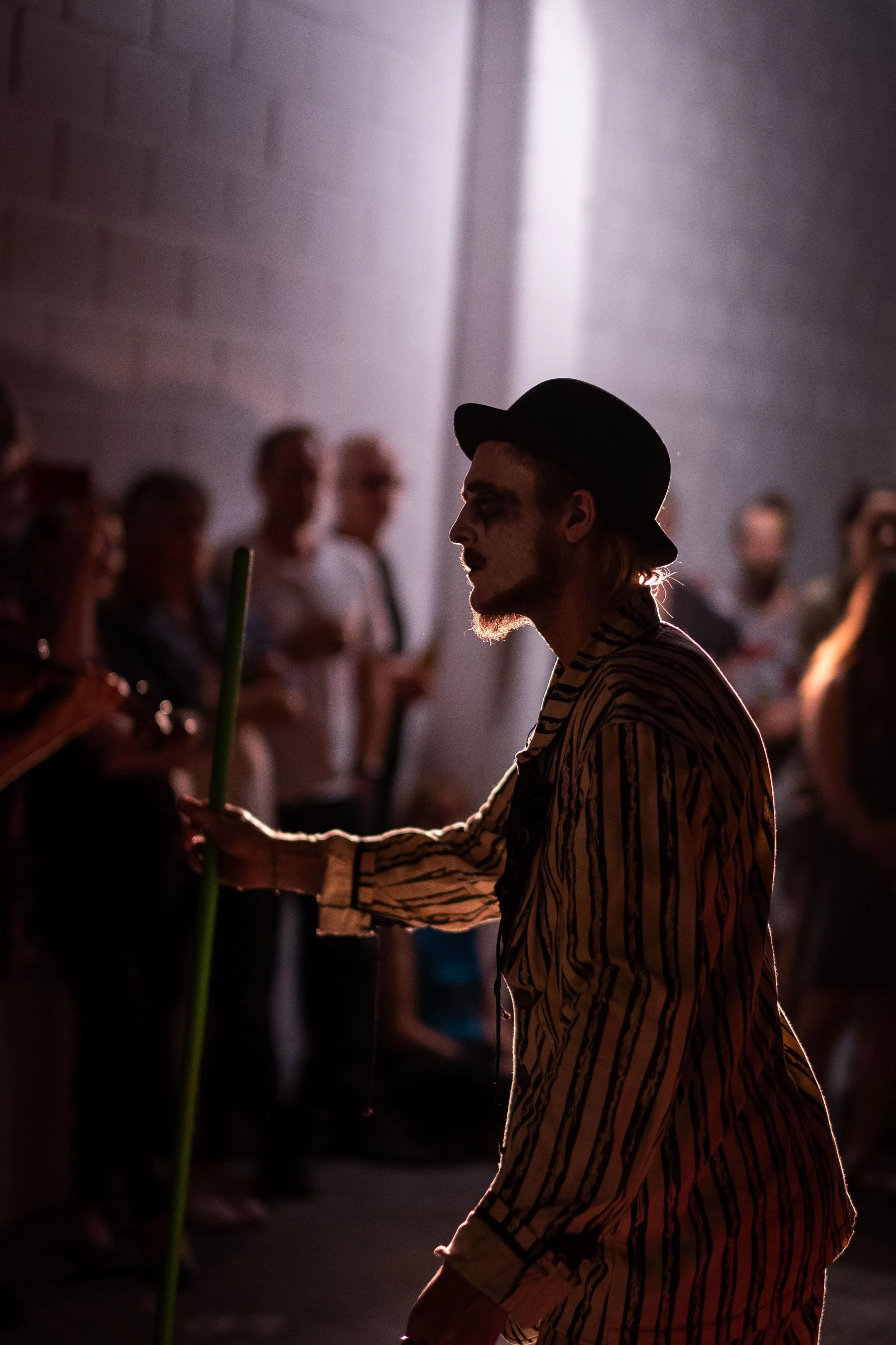 An actor dressed in a bowler hat, white and black pin stripped coat in a spotlight.  Around him in the dark is the audience observing the performance