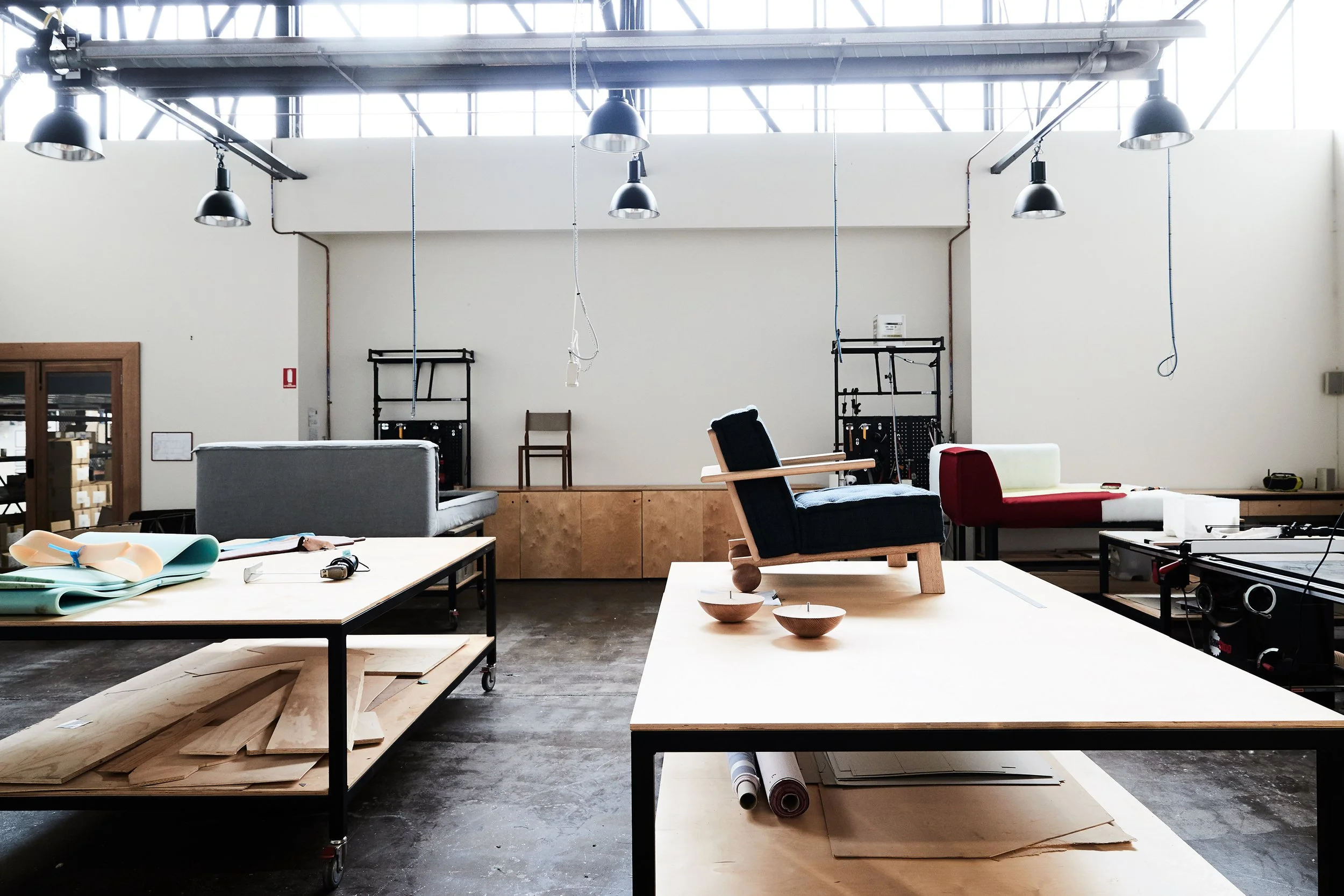 A spacious workshop or studio with chairs, wooden boards, and tools, illuminated by large overhead lights and natural light from high windows.