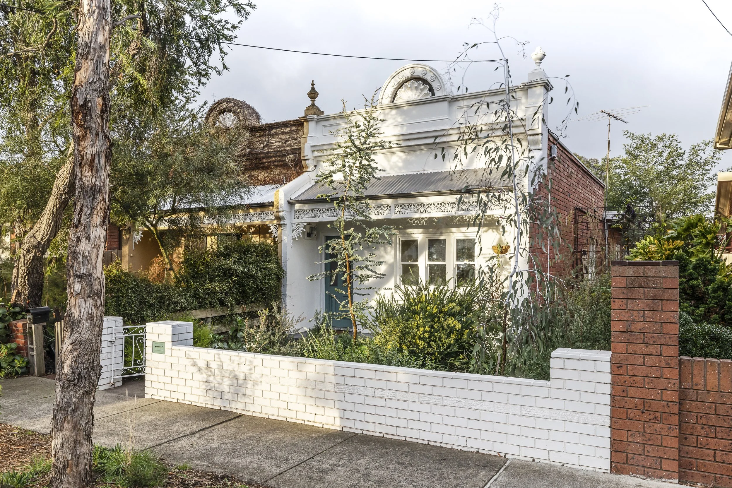 A house with a white facade, red brick wall, decorative architectural details, and a small front garden with trees and bushes, enclosed by a white brick wall and a red brick pillar.