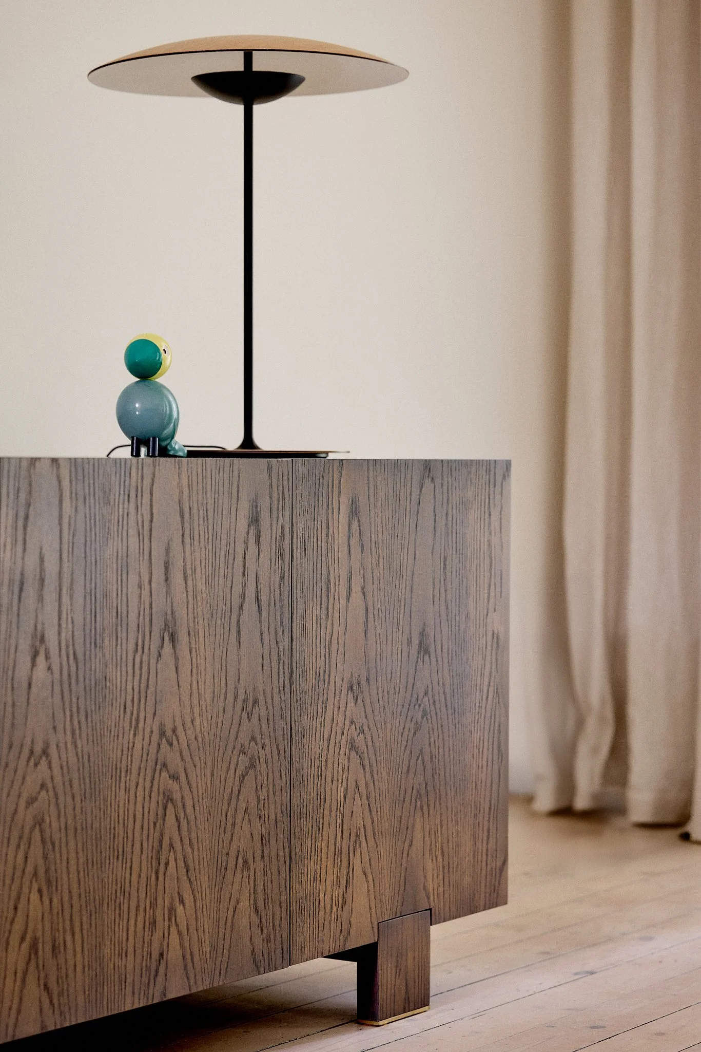 Close-up of a wooden cabinet with a small decorative figure and a modern table lamp on top, against a neutral background with curtains.