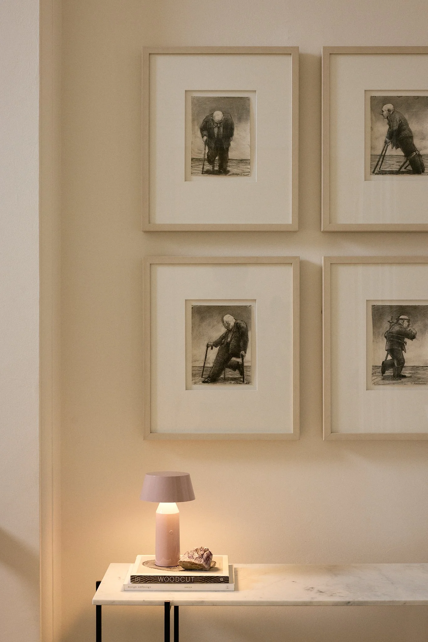 A decorated interior wall with four framed black-and-white drawings of an elderly man using a cane, spaced in a two-by-two grid. Below is a white marble console table with a small pink table lamp, a large rock, and a book titled "Woodcut."