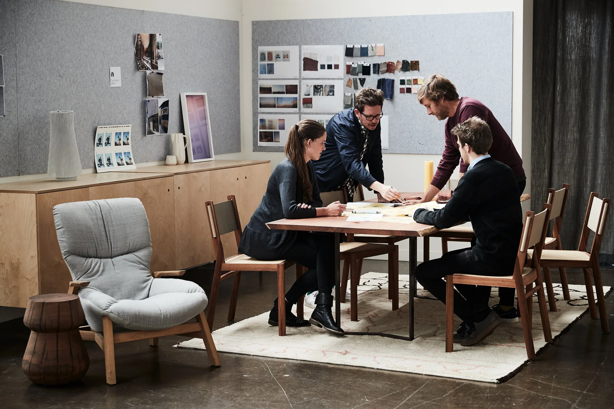 Four people gathered around a table in a modern office or conference room, reviewing documents and working together. The room has a gray textured wall with various images and design samples pinned up, and a light wooden sideboard with decorative item