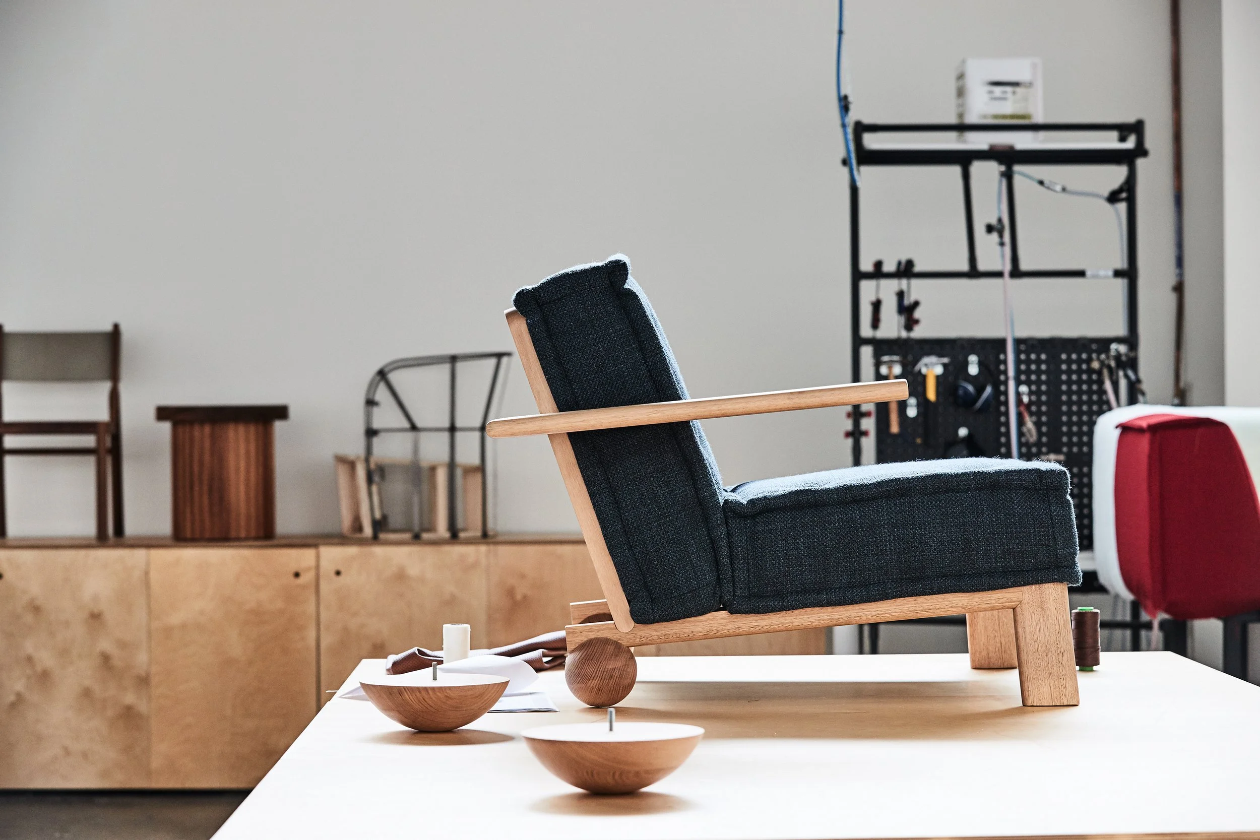 A modern armchair with a wooden frame and black cushioned seat, placed on a white table in a furniture store. In the background, various chairs and furniture pieces are visible, along with a black metal shelving unit with tools and equipment.