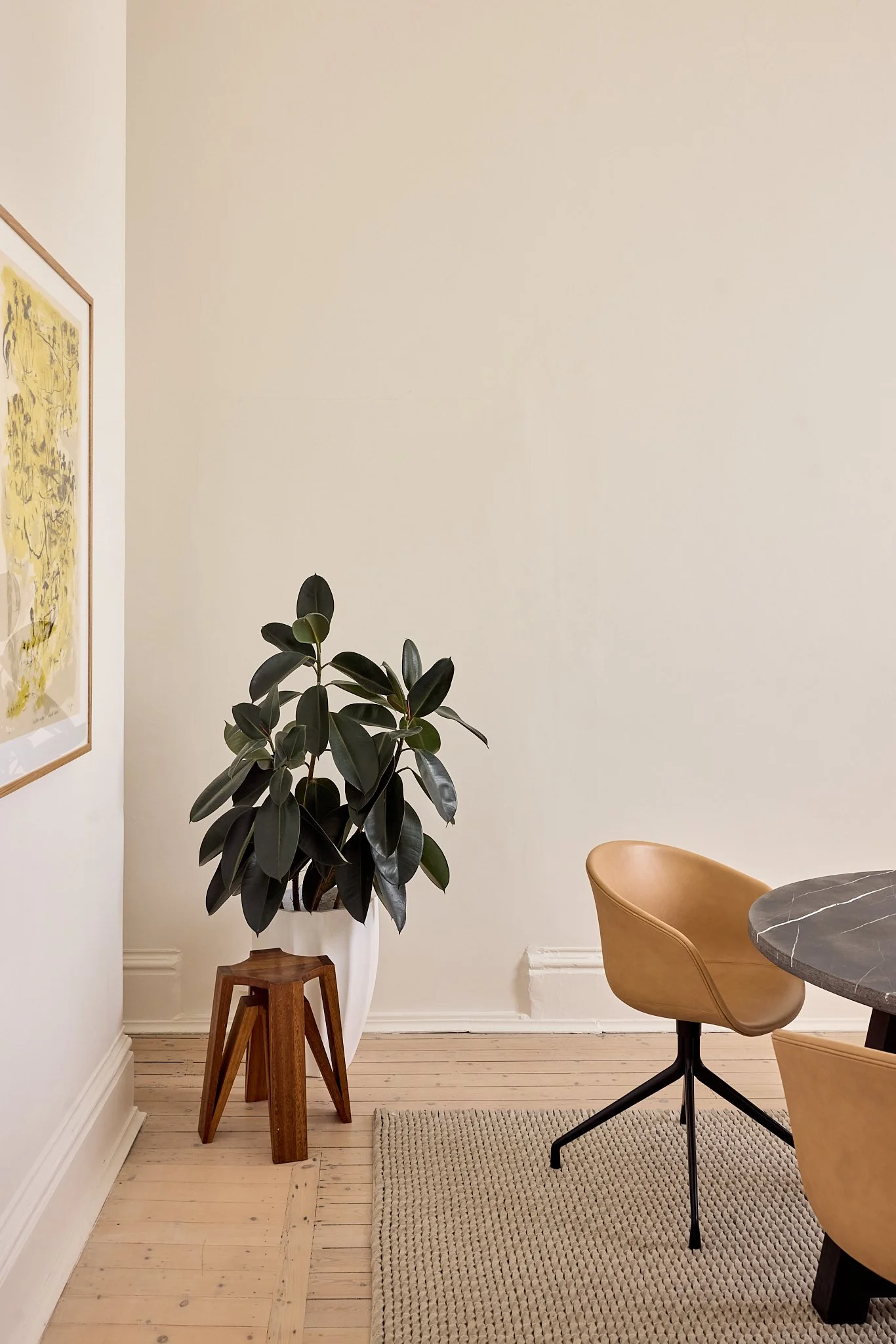 Interior room corner with a large potted plant on a wooden stand, a table with a marble top, and two tan chairs.