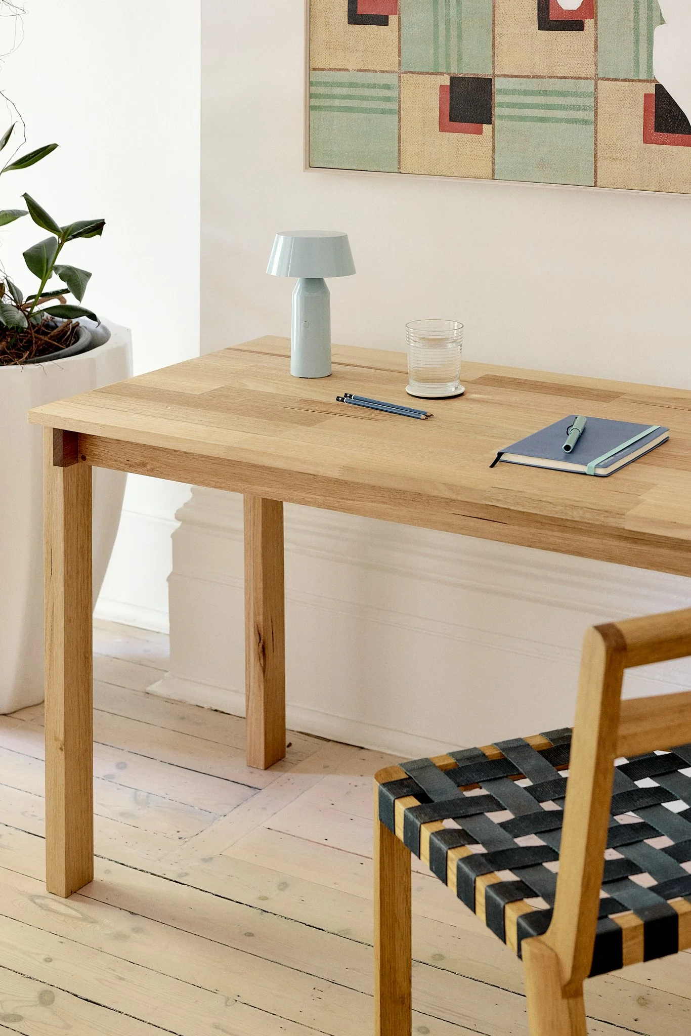 A minimalist wooden desk with a blue notebook, a pen, a small table lamp, a glass of water, and a plant in a white pot nearby. A modern chair with a woven black and natural strap seat is partially visible in the foreground, and a colorful geometric art piece hangs on the wall behind.