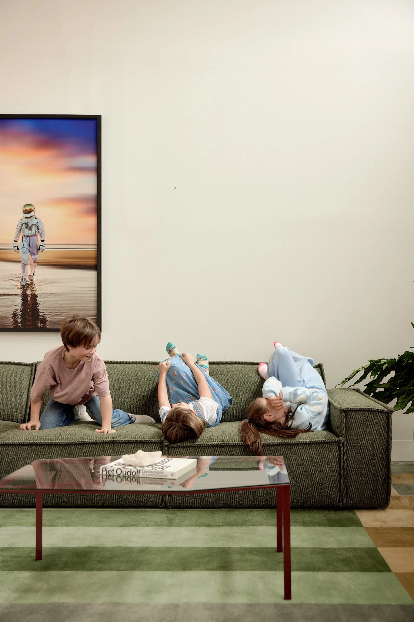 Three children are playing and relaxing on a green sofa in a living room. One girl is lying down on her back, another girl is sitting near her, and a boy is on his hands and knees. There is a glass-top coffee table in front of them with books and a t