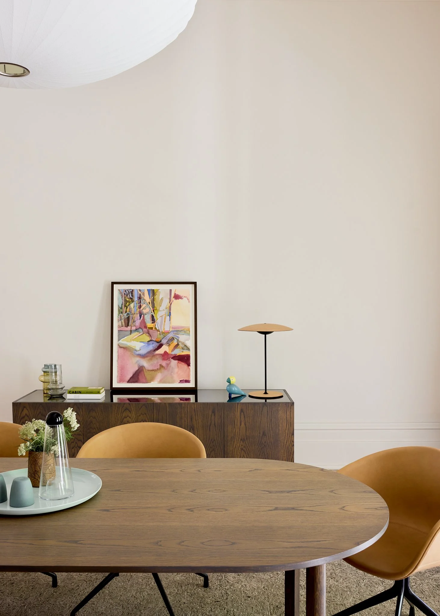 Minimalist dining room with a wooden table, tan chairs, a sideboard with decorative items, a colorful abstract painting, a small lamp, a duck figurine, and a vase with white flowers.