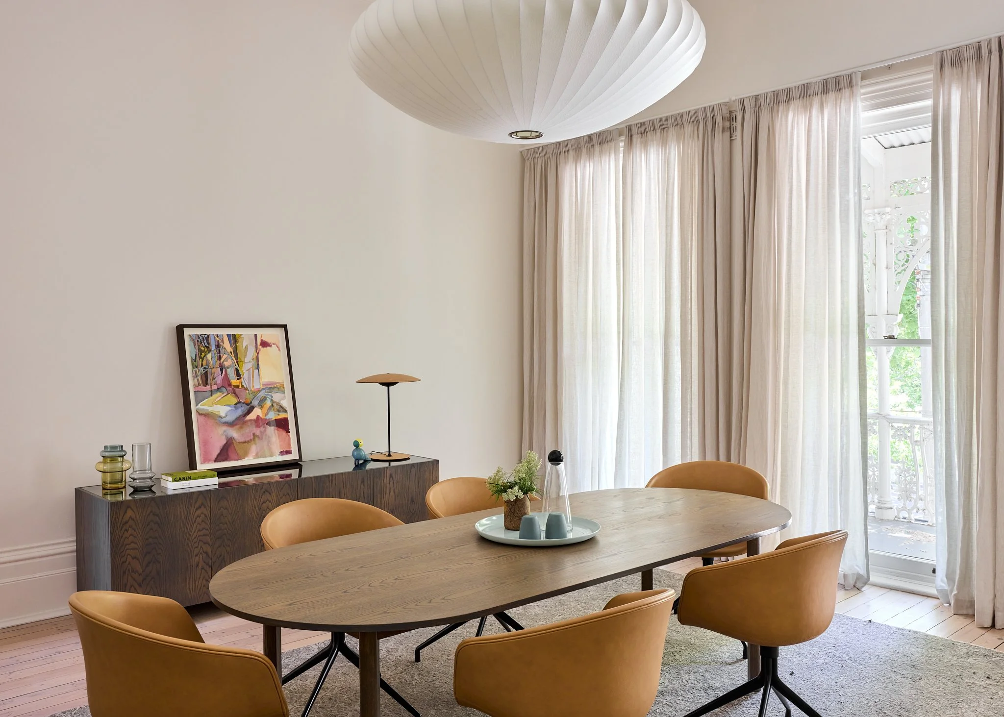 Modern dining room with a wooden oval table, six tan chairs, a sideboard with decorative objects, a painting, and a large window with sheer curtains letting in natural light.