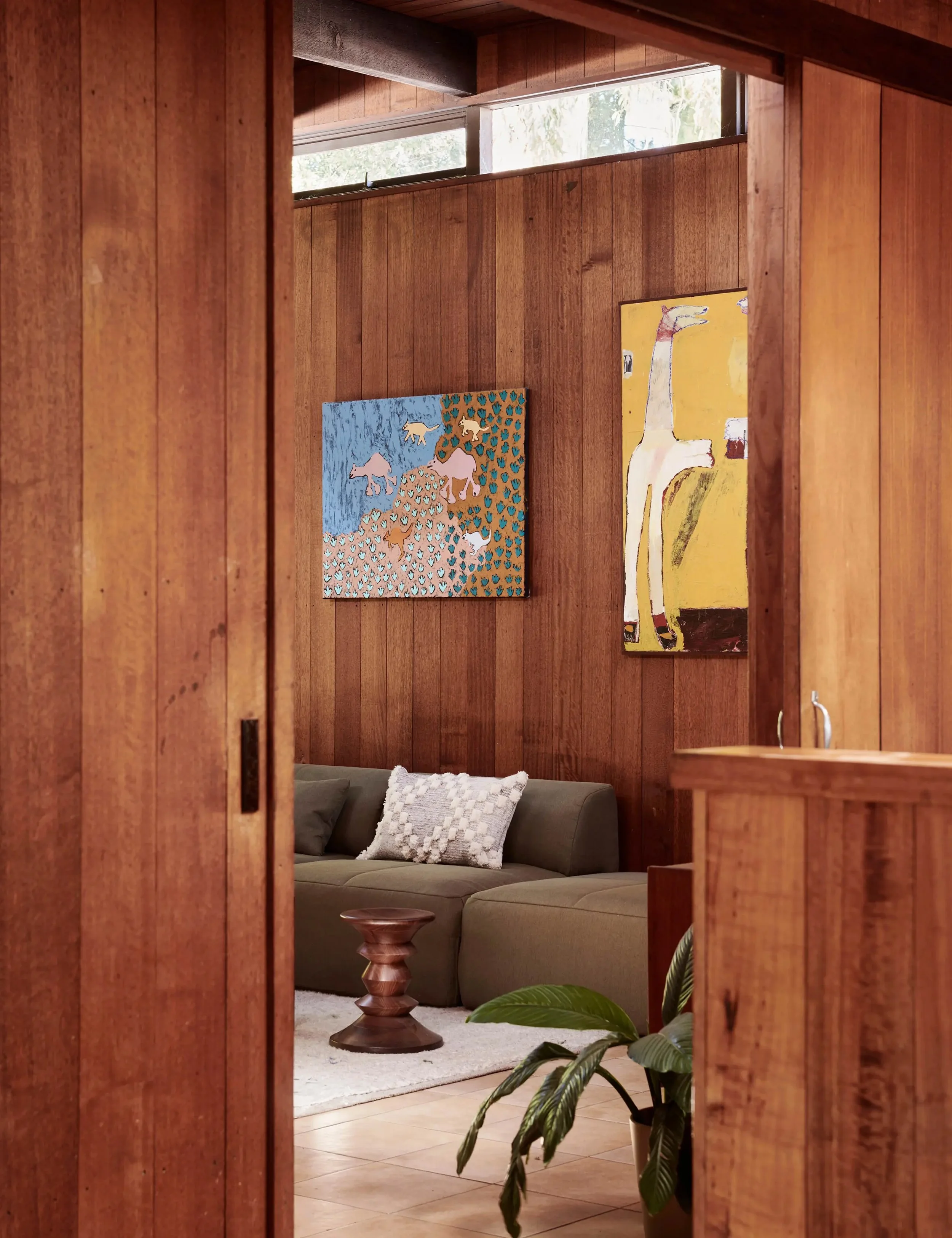 View into a living room through a partially open wooden door, showing a green couch with a decorative pillow, a small wooden side table, colorful animal-themed paintings on the wooden wall, and a potted plant.