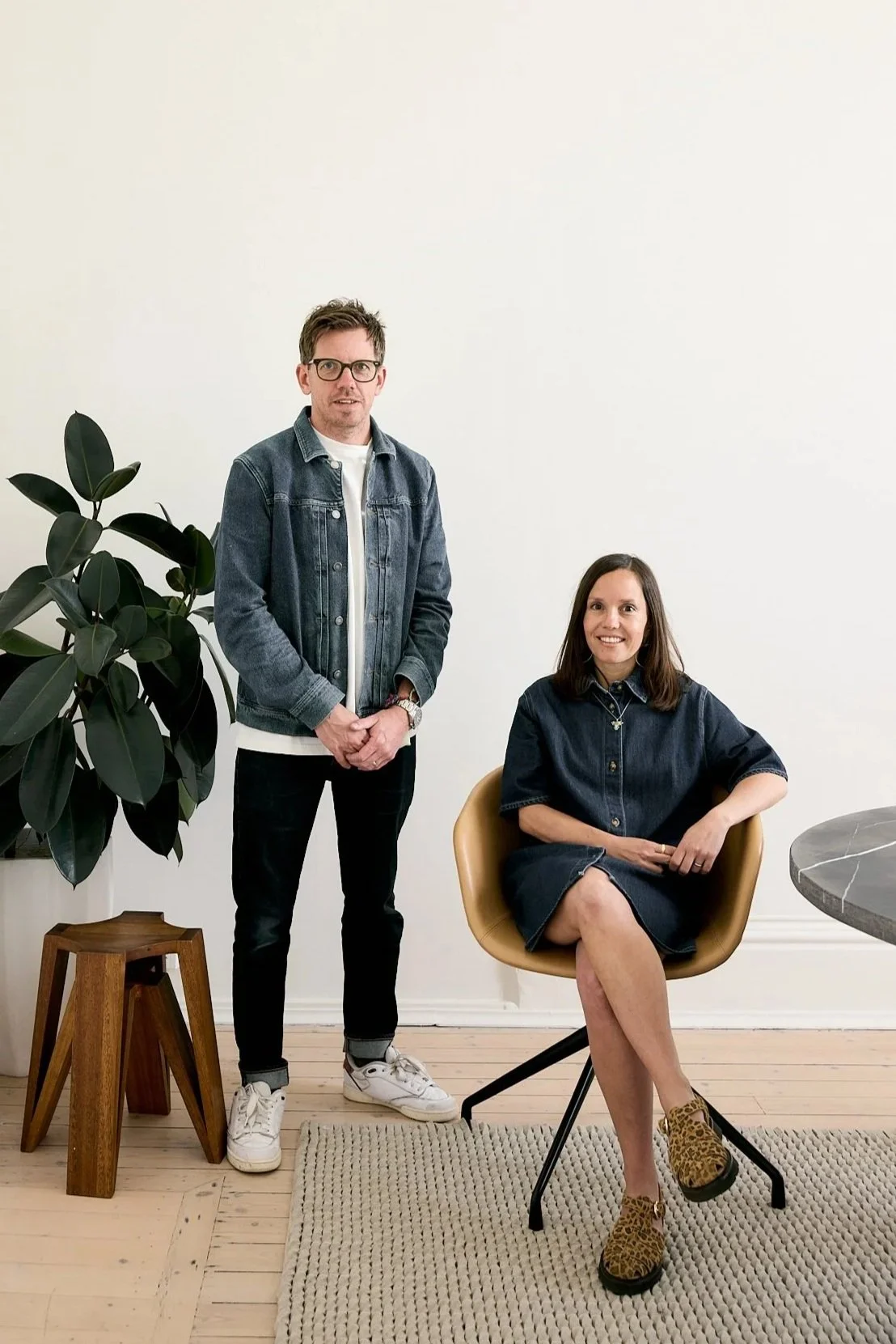 A man and woman with light skin posing indoors against a plain white wall, with a large potted plant on a wooden stand to their left. The man is standing, wearing glasses, a denim jacket, white shirt, black pants, and white sneakers. The woman is seated on a tan chair, wearing a dark denim dress with rolled-up sleeves and leopard print shoes, smiling at the camera.