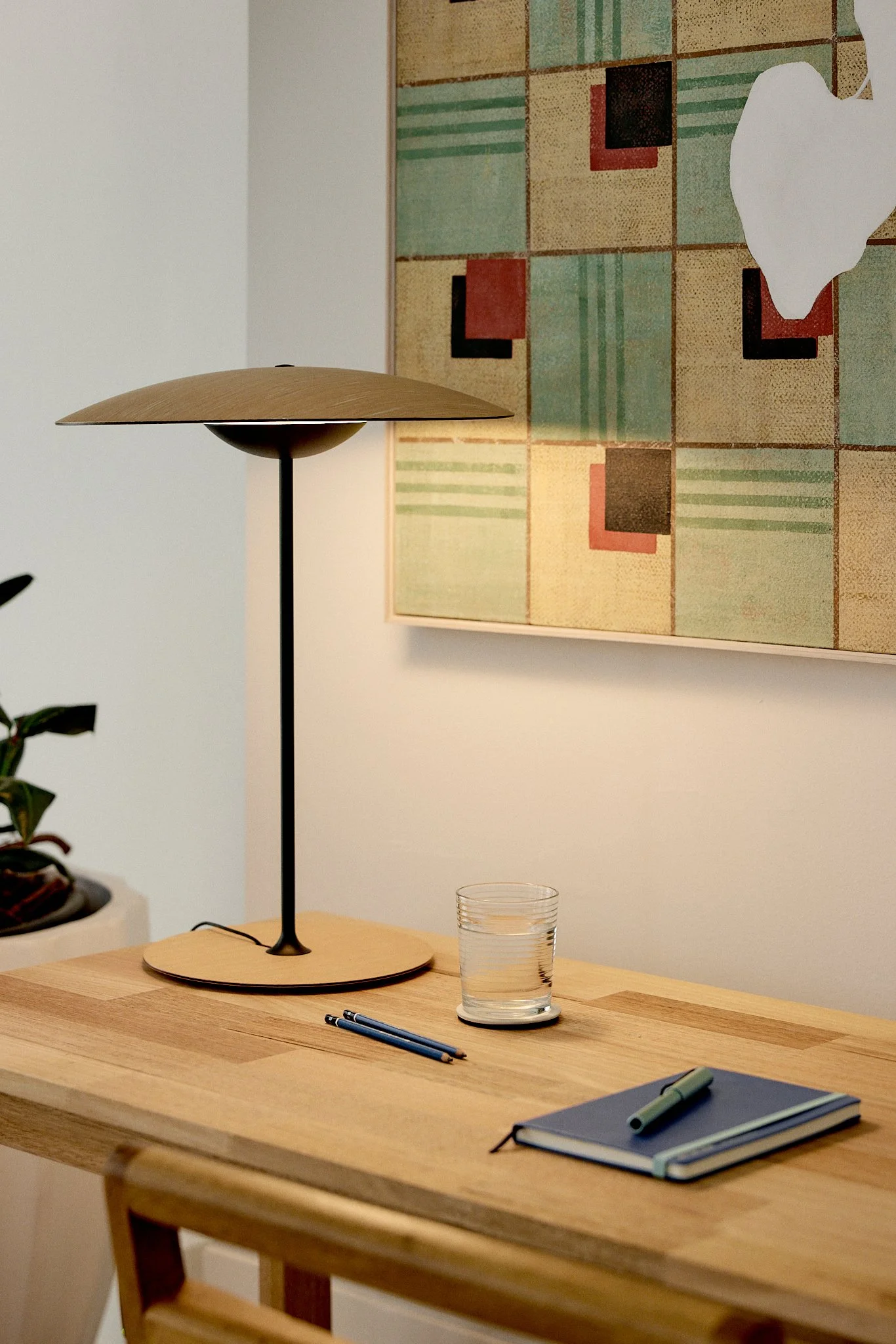 A wooden desk with a modern lamp, a glass of water, a closed notebook, two pens, and a small plant in the background, with abstract artwork on the wall.