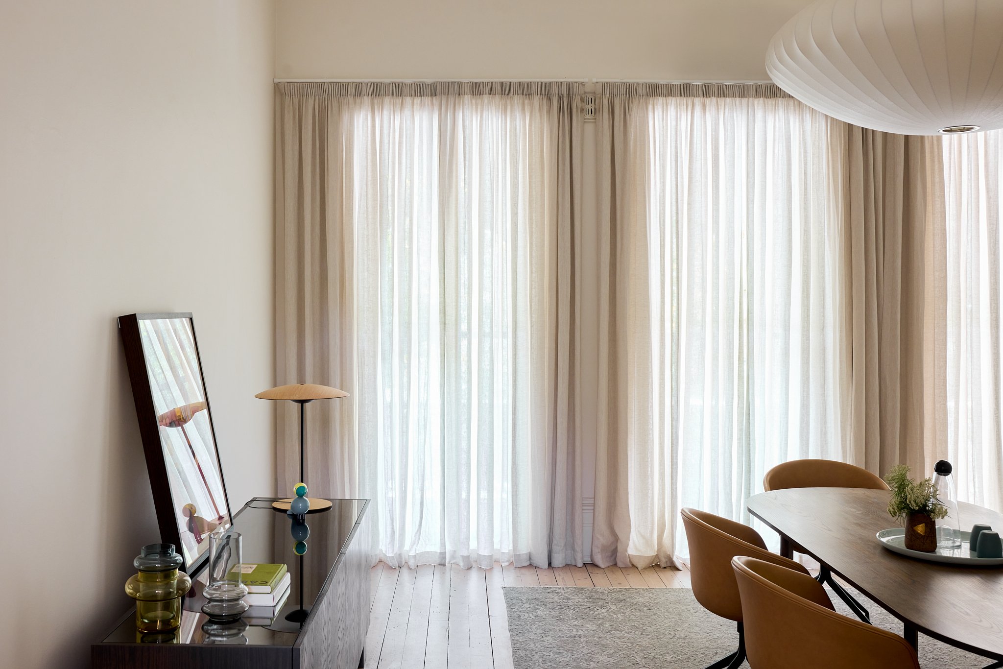 Living room with sheer white curtains covering large windows, wooden dining table with yellow chairs, sideboard with mirror, table lamp, decorative objects, and a potted plant