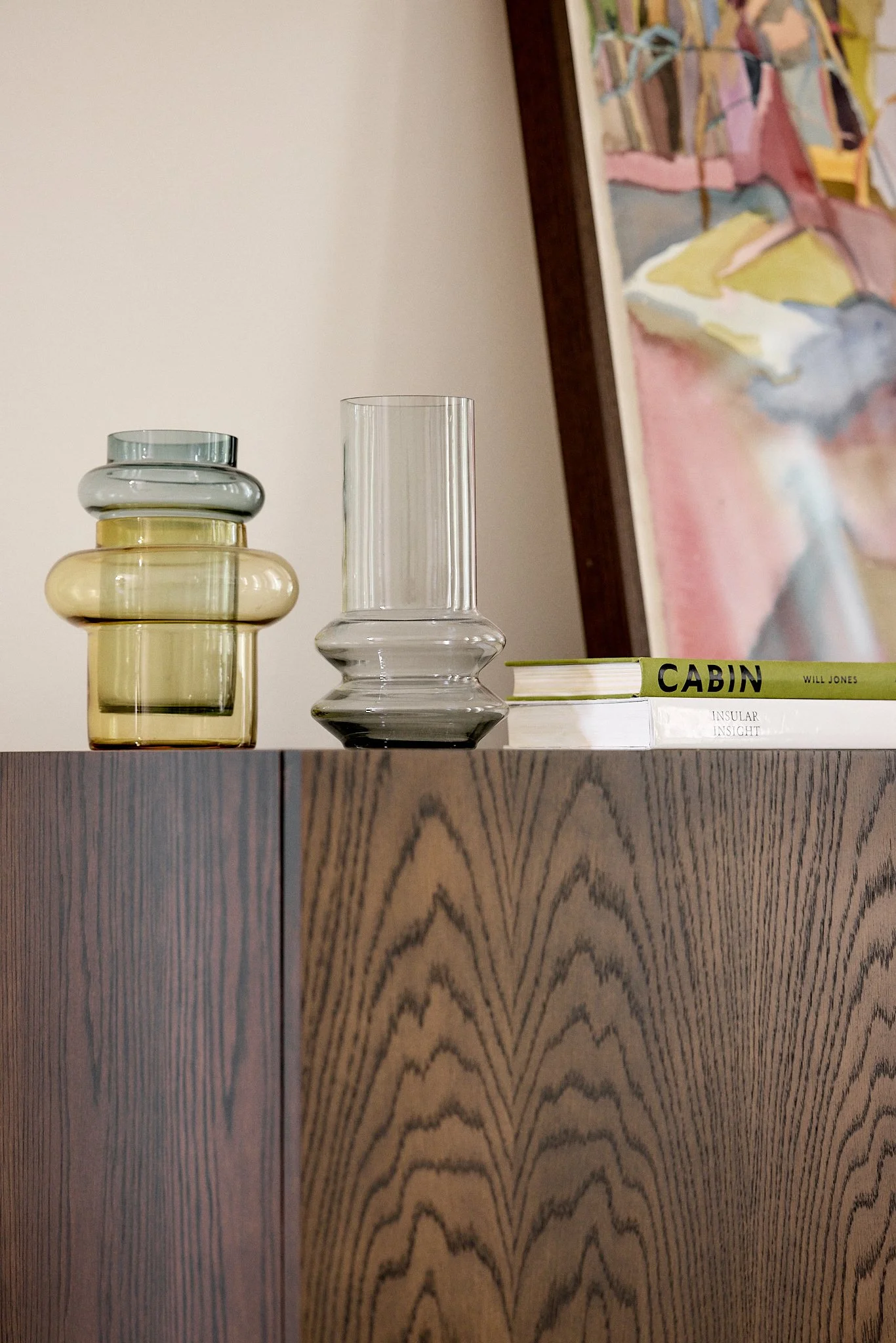 Decorative glass vases and books on top of a wooden cabinet, with a framed artwork in the background.