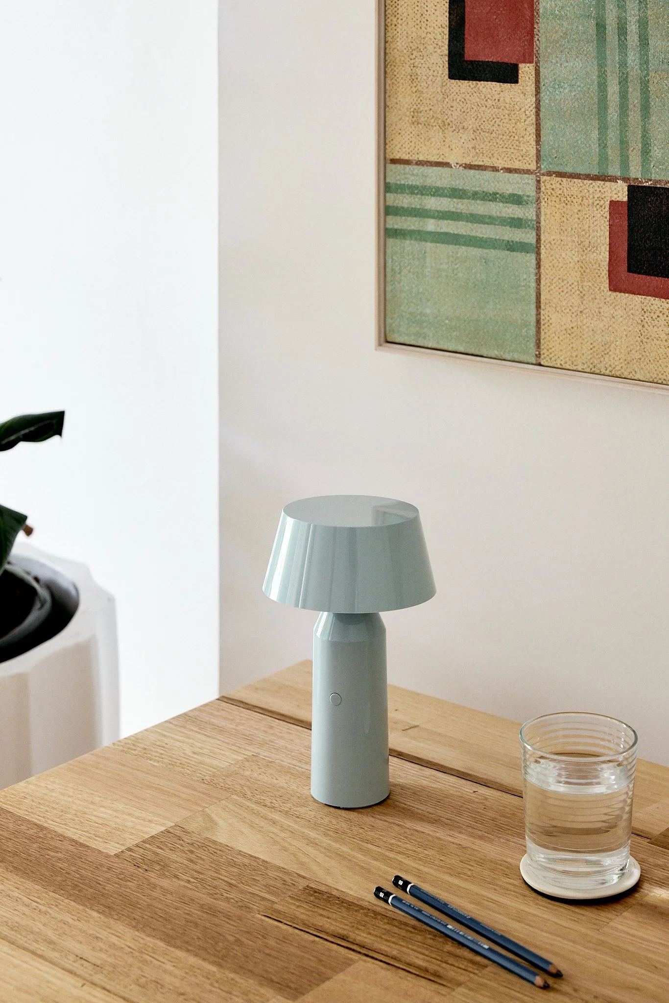 A modern table lamp with a gray base and shade on a wooden table next to a glass of water and two pencils, with a framed abstract artwork on the wall behind.