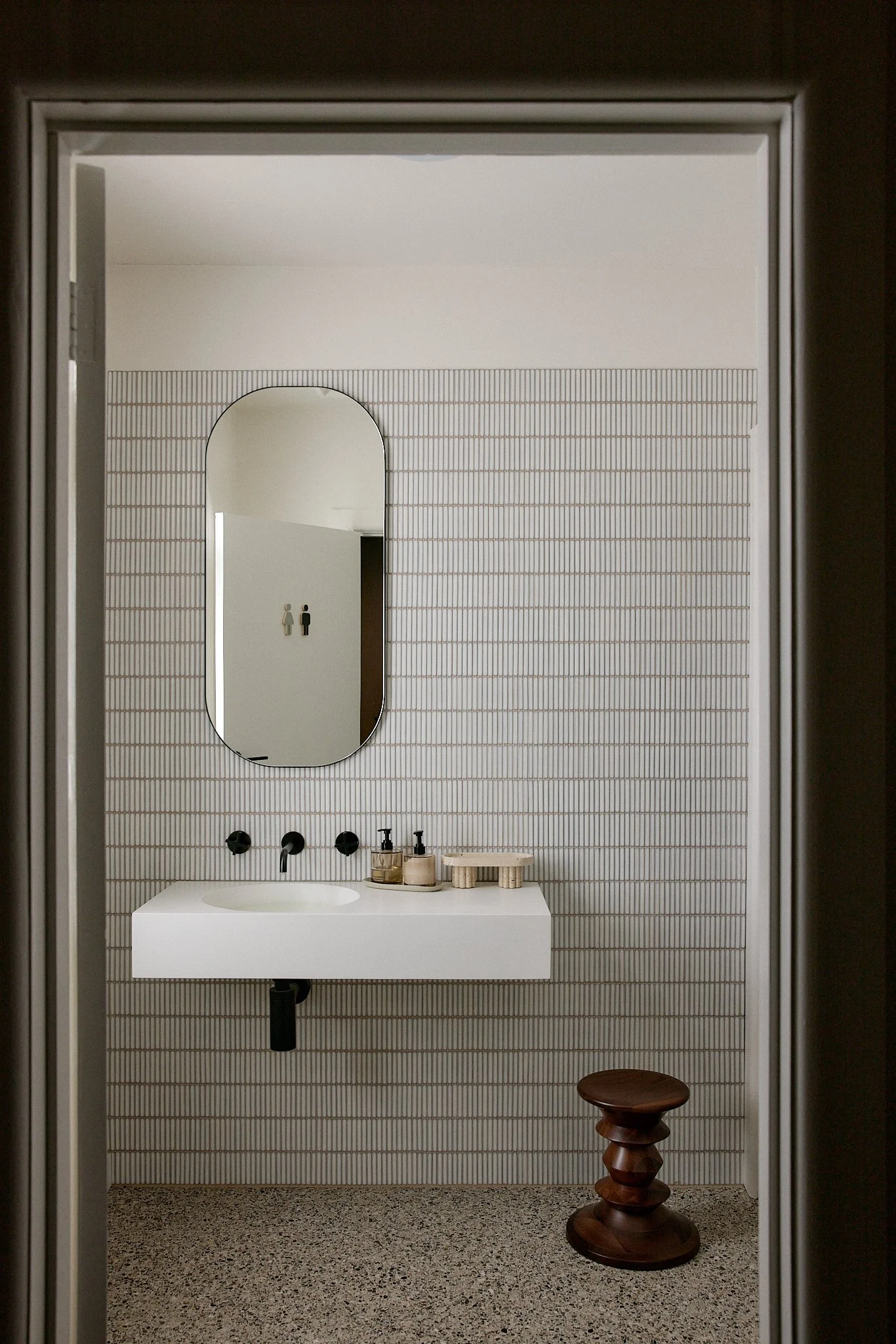 Minimalist bathroom with a white rectangular sink, round mirror, black fixtures, wooden stool, and small accessories on the sink.