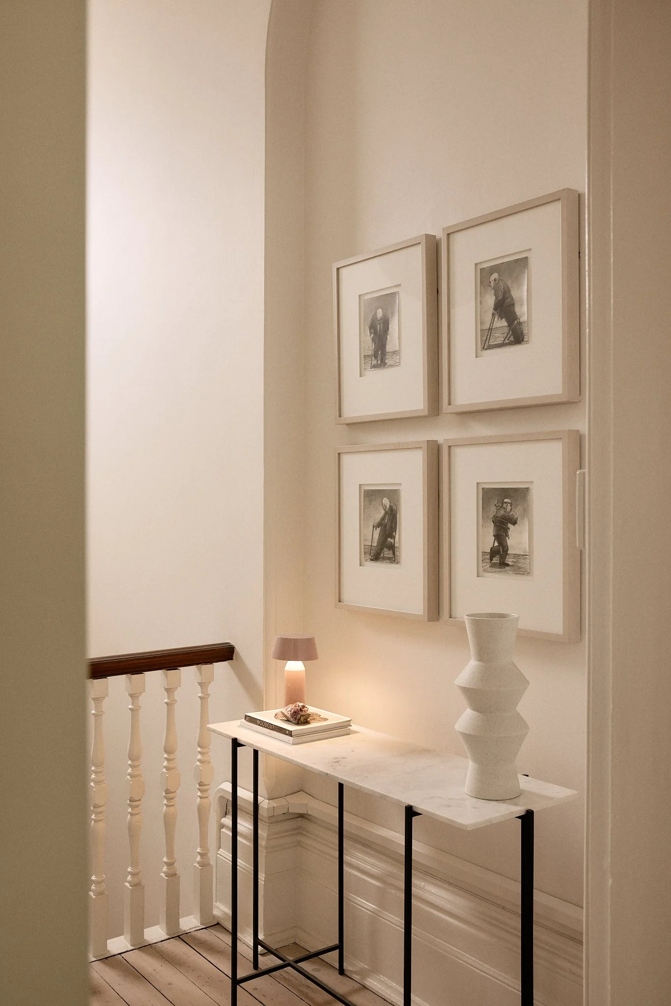 A hallway corner with four black-and-white vintage photographs of elderly men, a small table with a pink lamp, books, a shell, and a white upcoming sculpture, against beige walls with a wooden railing.