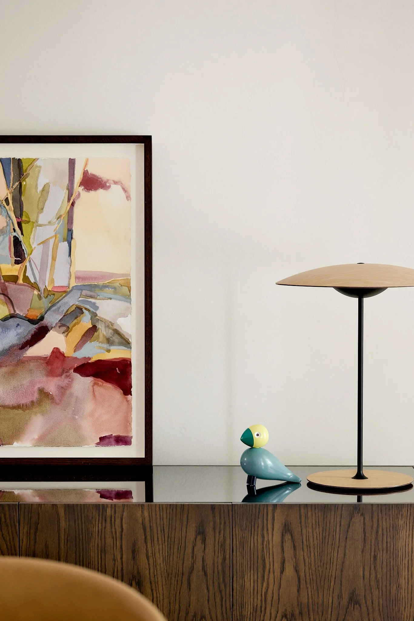 A wooden sideboard with an abstract watercolor painting, a modern table lamp with a wooden shade, and a small decorative bird figure on a white wall background.