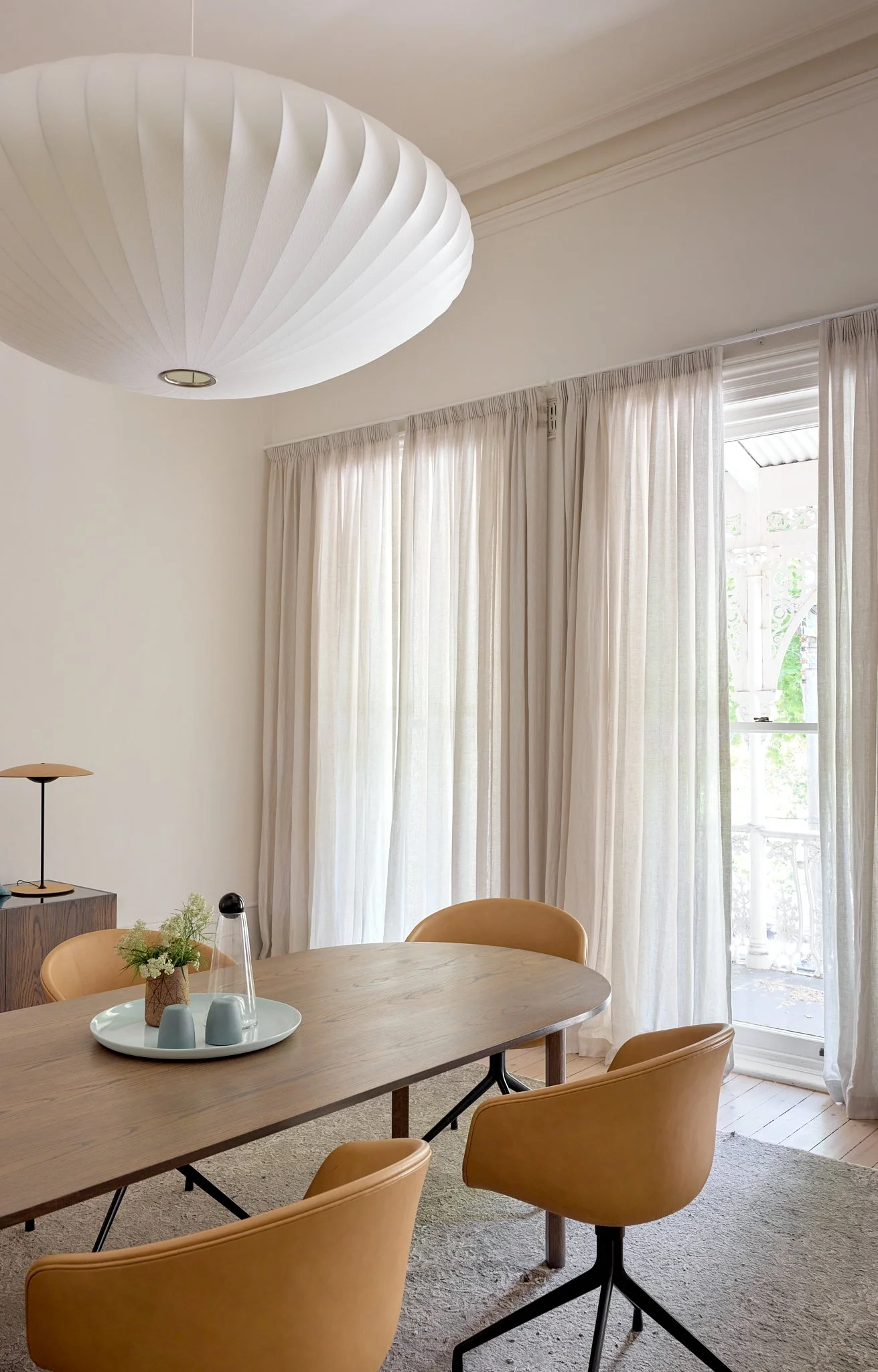 A dining room with a wooden table, tan chairs, sheer white curtains, and a large white pendant light fixture.