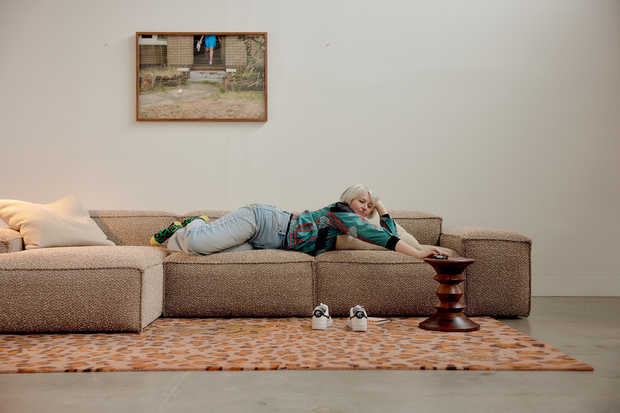 An elderly woman with white hair lying on her stomach on a beige sectional sofa in a room with white walls. She is wearing light-colored pants and a colorful patterned shirt, reaching towards a dark wooden side table. There are sneakers on the floor 
