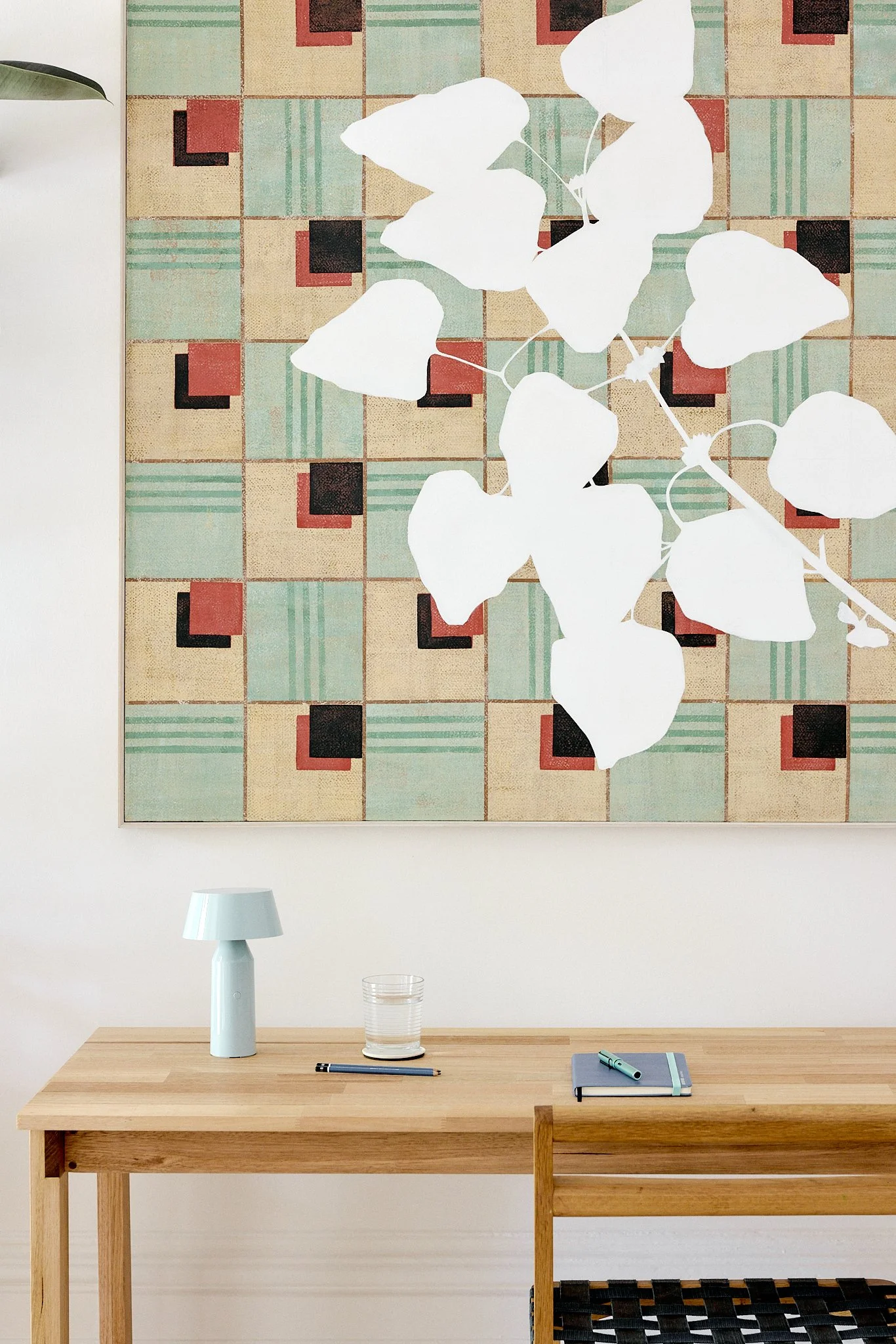 A minimalist wooden desk with a small light blue lamp, a glass of water, a closed notebook, and a pen, in front of a wall with a colorful, geometric patterned artwork. There is also a chair with a woven seat partially visible.
