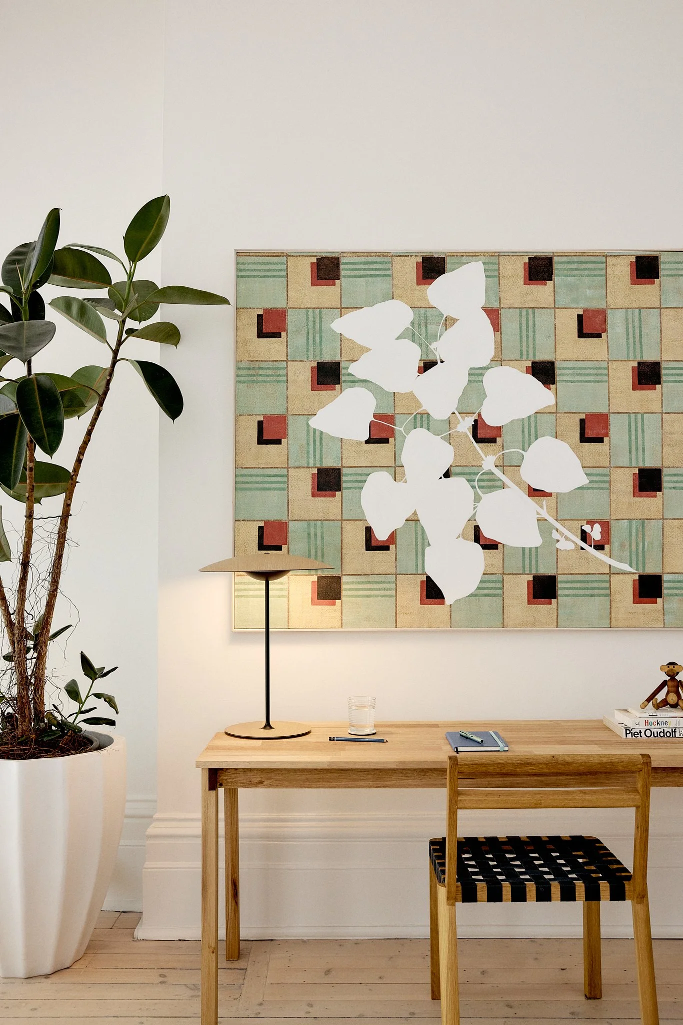 Interior scene with a large potted plant, a wooden desk with a table lamp, a glass of water, and a small notebook, a wooden chair with a woven seat, and a colorful geometric artwork on the wall.