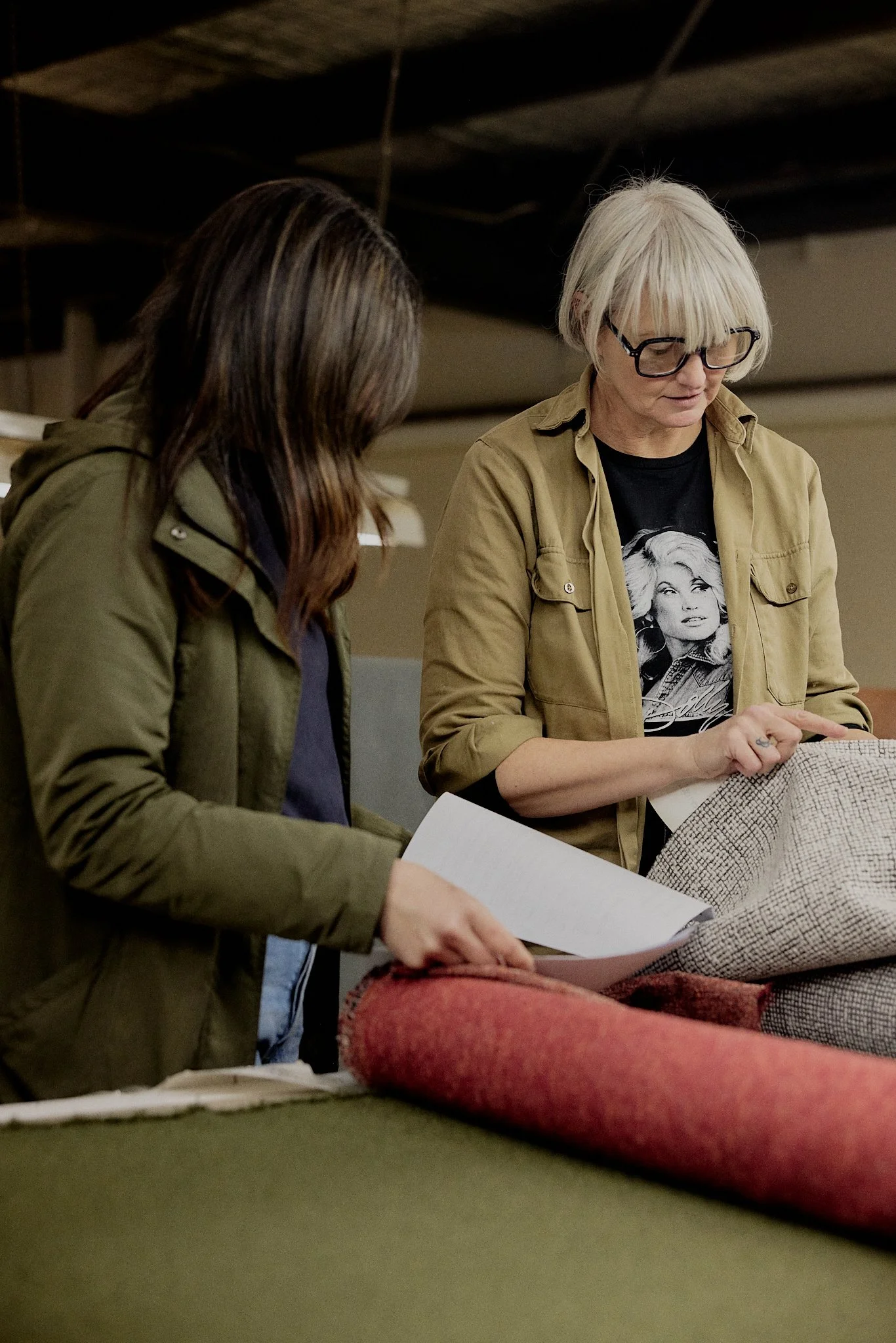 Two women examining fabric samples or design plans in a workshop or showroom, with rolls of fabric in front of them.