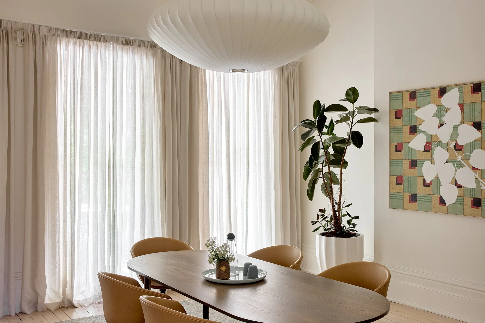 A modern dining room with a wooden table and six tan chairs, a large leafy plant in a white pot, sheer curtains over large windows, and an abstract geometric painting on the wall.