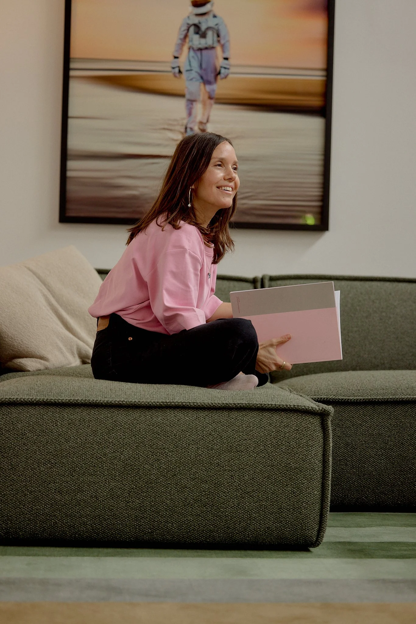 A woman with shoulder-length brown hair, wearing a pink shirt and black pants, sitting on a green couch holding a pink and gray folder, smiling, with a large framed photograph of an astronaut walking on the surface of a planet or moon in the backgrou