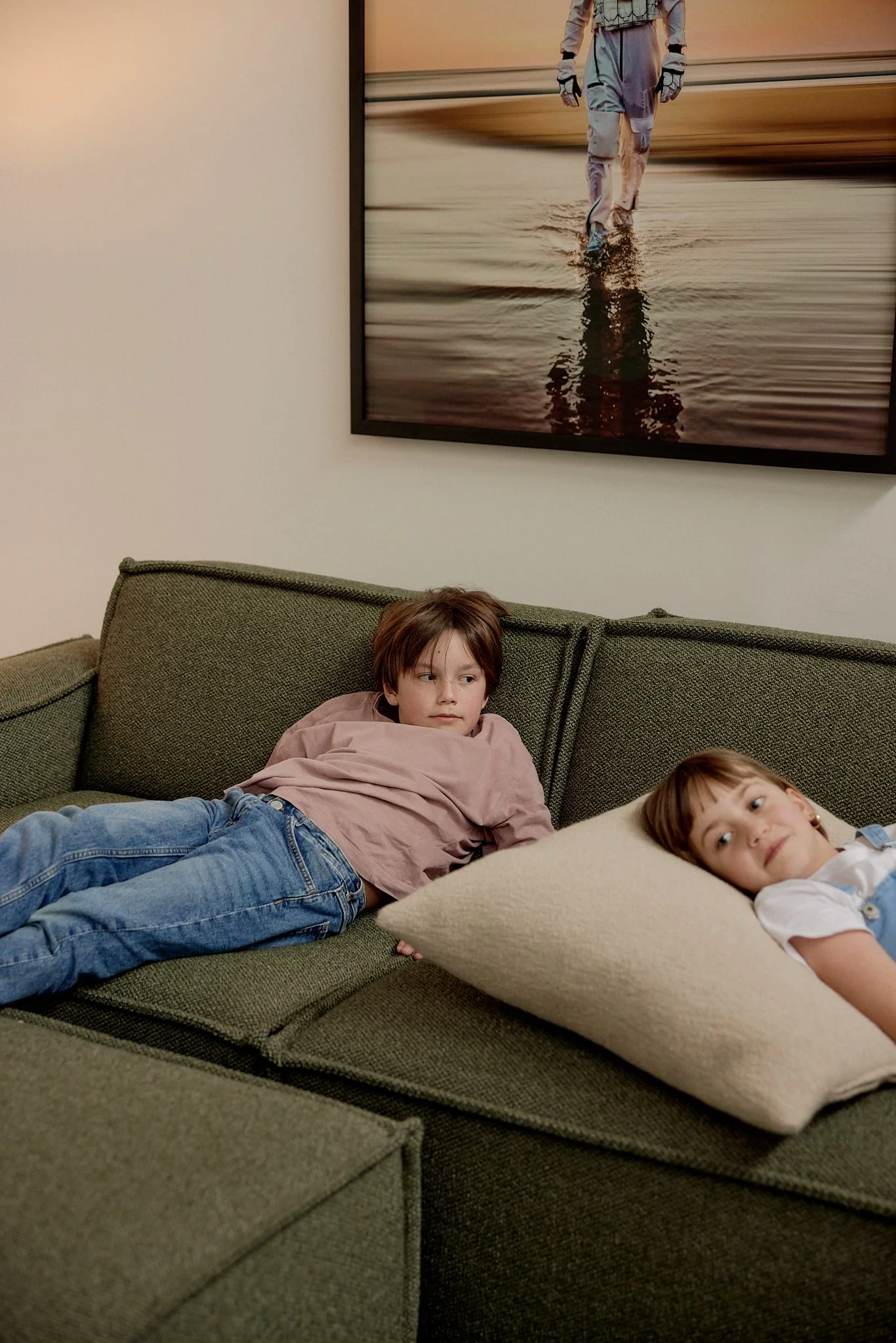 Two children lying on a green couch in a living room, one with brown hair and the other with light brown hair, resting on a pillow, with a large framed photo of a person walking on water in the background.