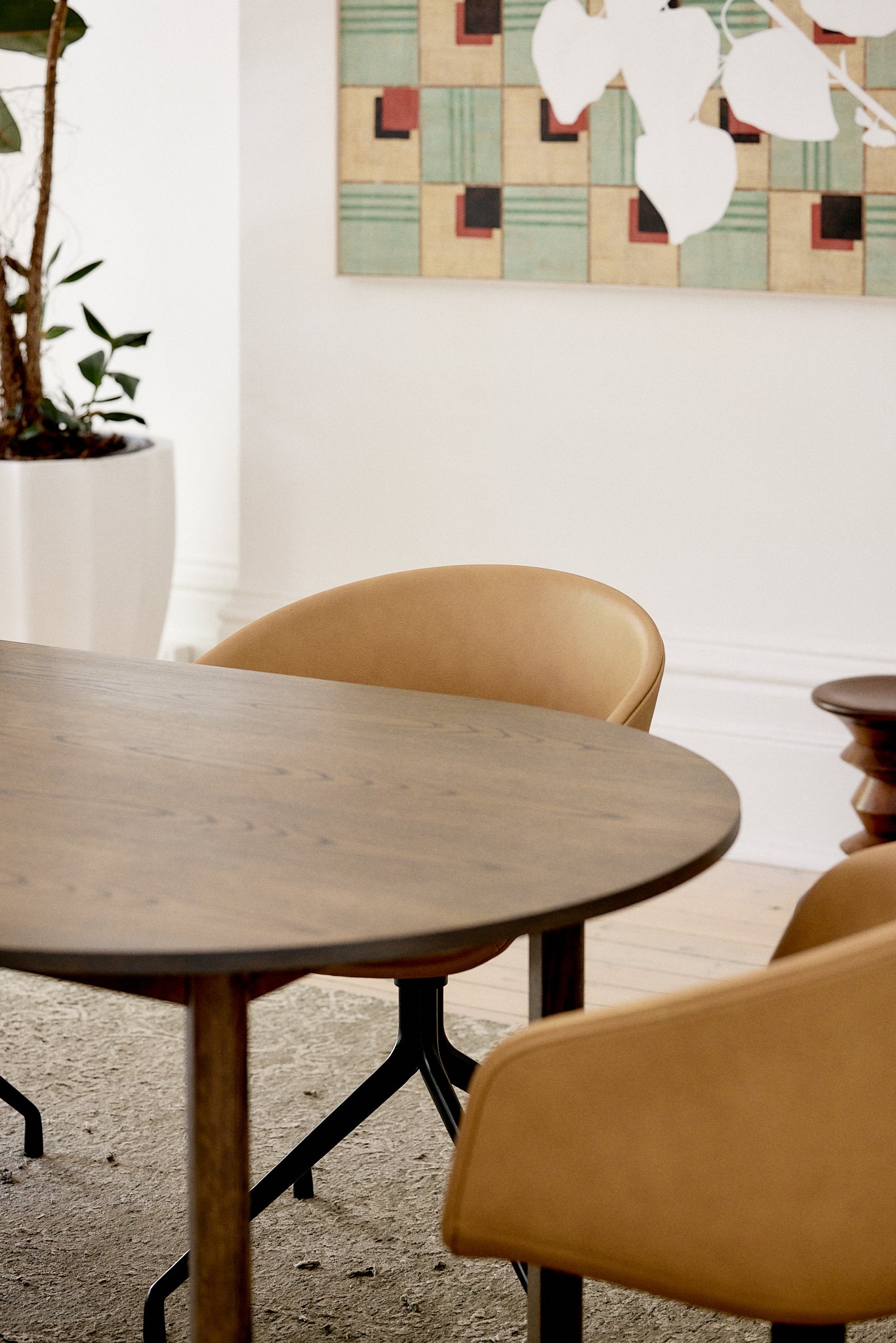 A round wooden table surrounded by tan chairs in a modern room with a large potted plant, wall art, and a textured beige rug.