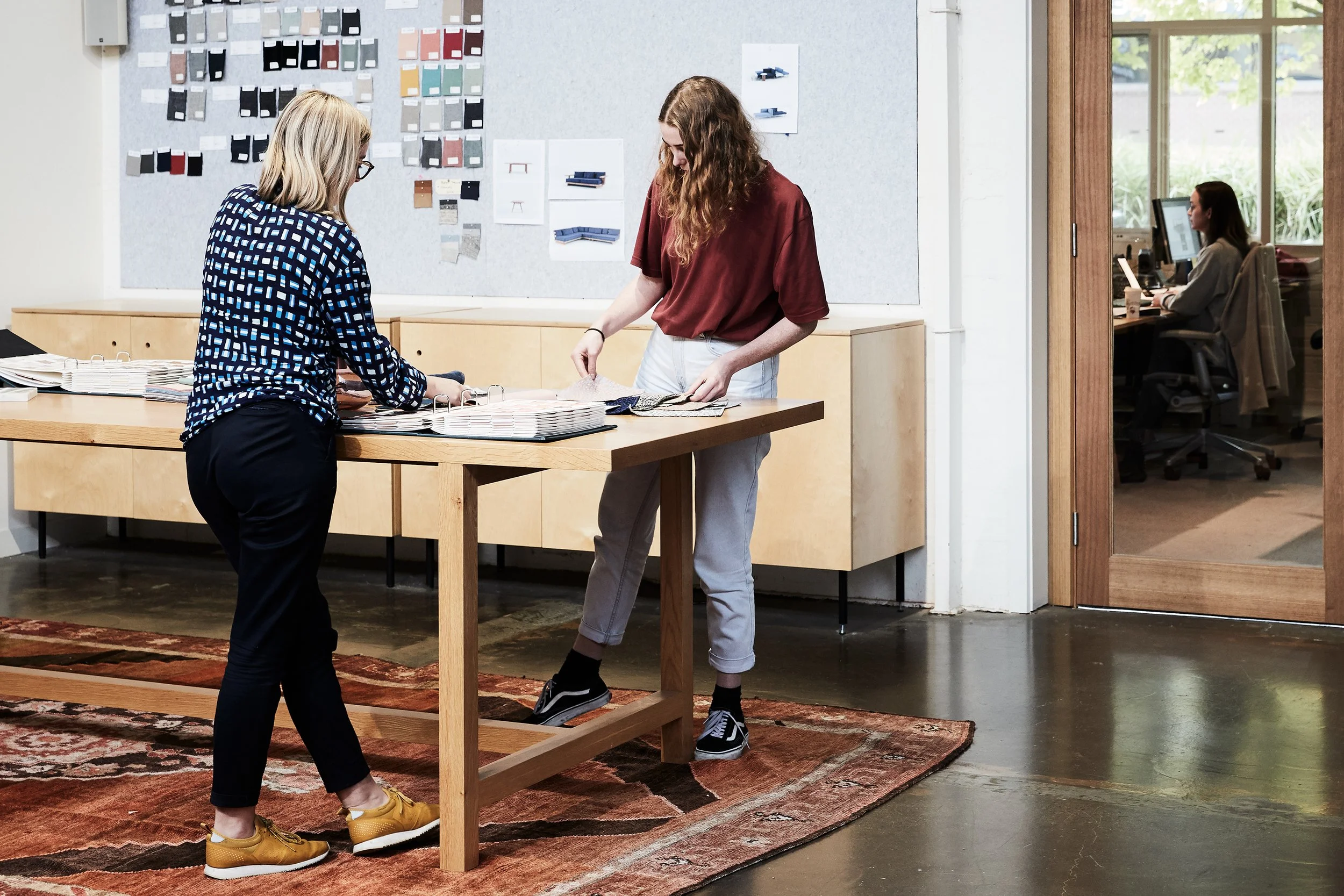 Two women standing at a wooden table reviewing fabric samples, with color swatches and design plans on the wall behind them, in a modern office with a glass door leading to an employee working at a desk.