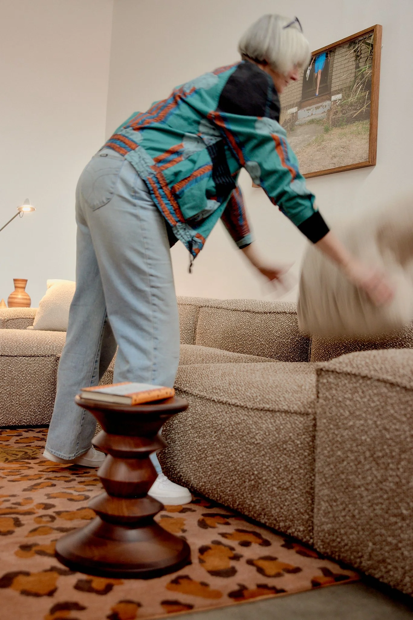 Person with silver hair in a colorful jacket and light-colored pants, bending over to pick up a pillow on a beige sofa in a living room with a patterned carpet and a small wooden side table.