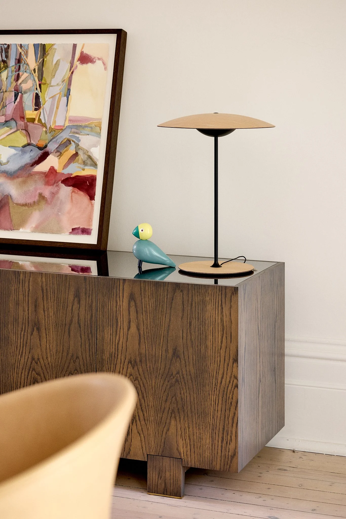 A wooden cabinet with a glass top, decorated with a colorful abstract painting, a modern bird-shaped figurine, and a minimalist table lamp.