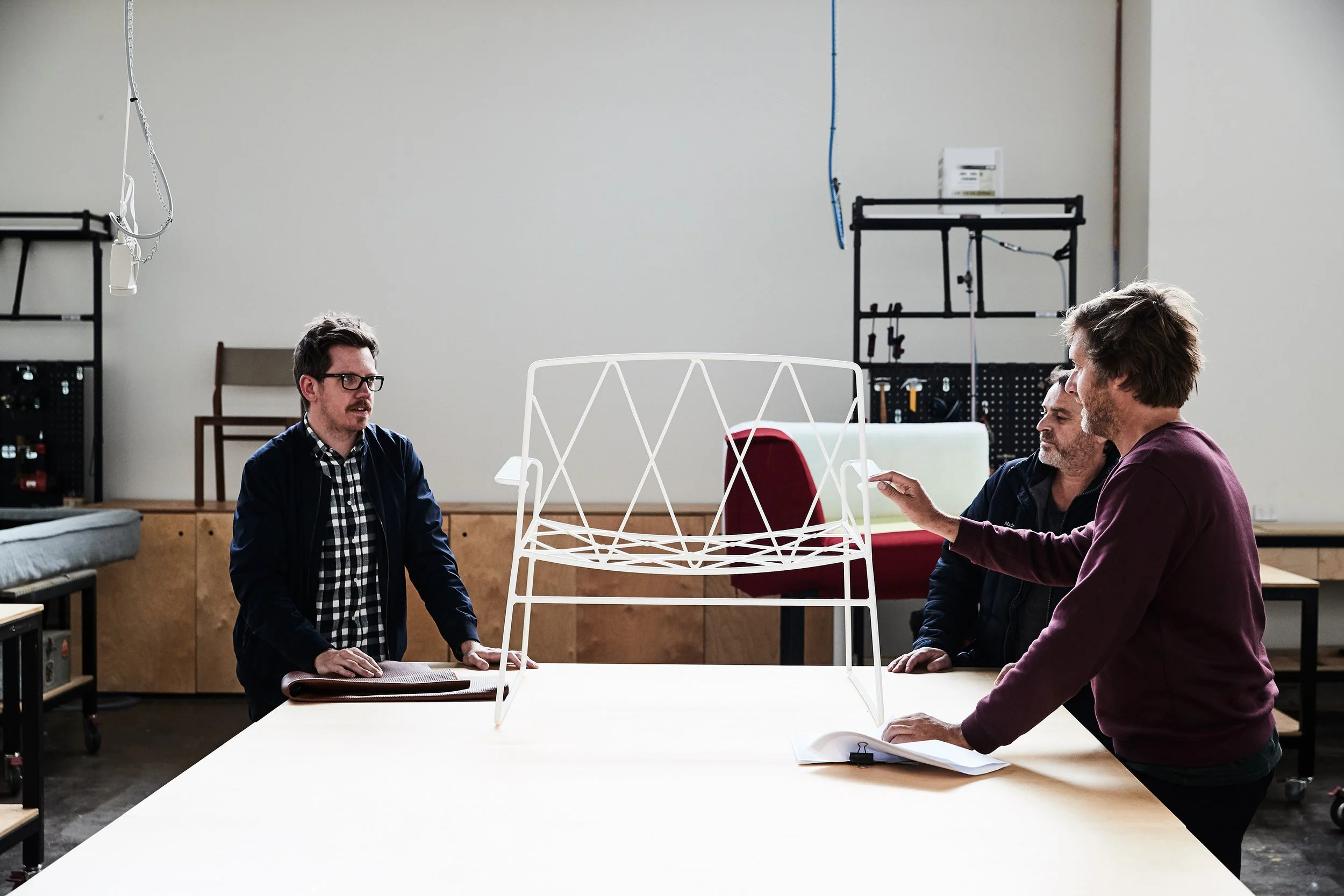 Three men discuss a wireframe sculpture of a chair in a workshop.