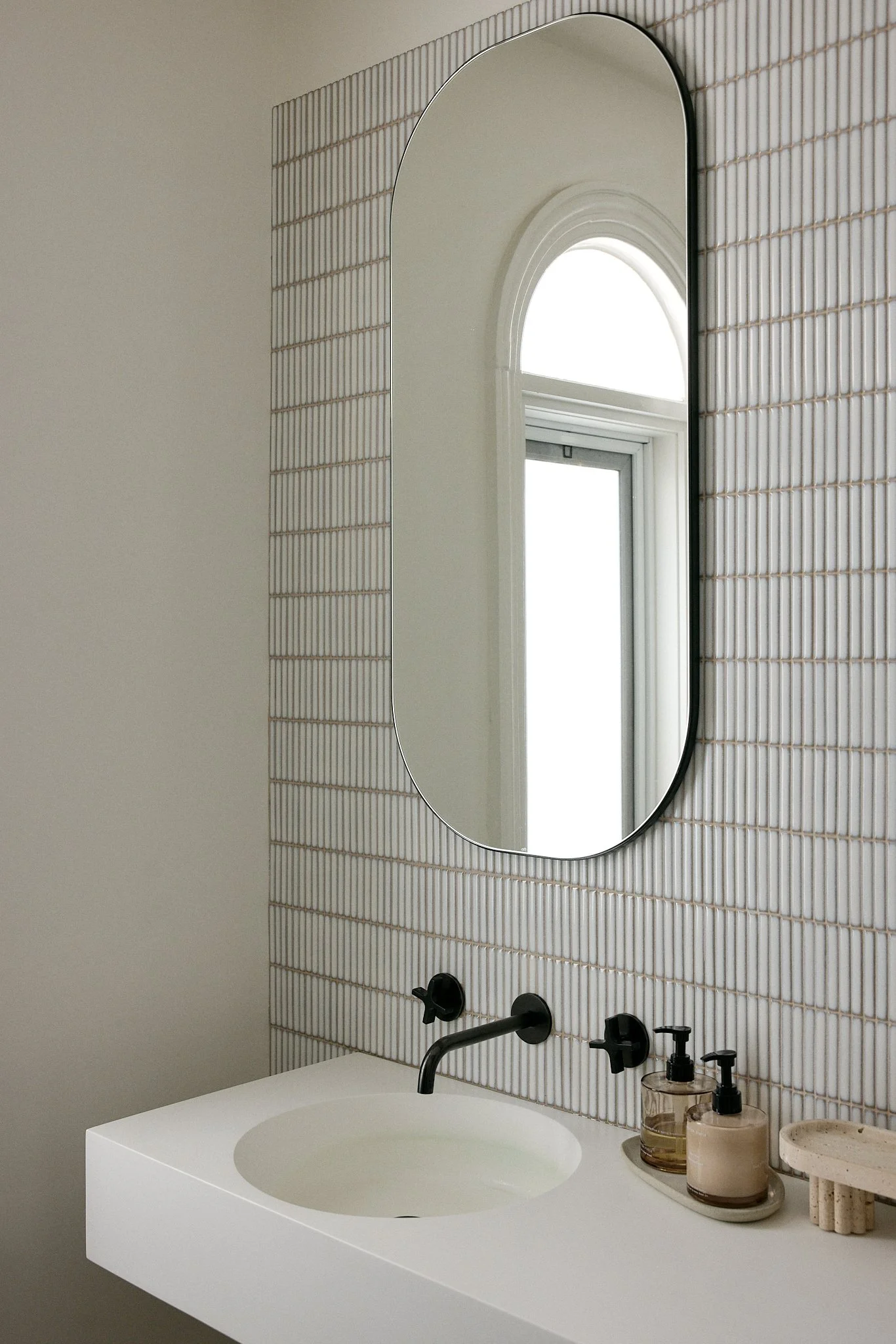 Minimalist bathroom sink area with a large oval mirror, black faucet, and modern soap dispensers, reflected in a window with a rounded arch.