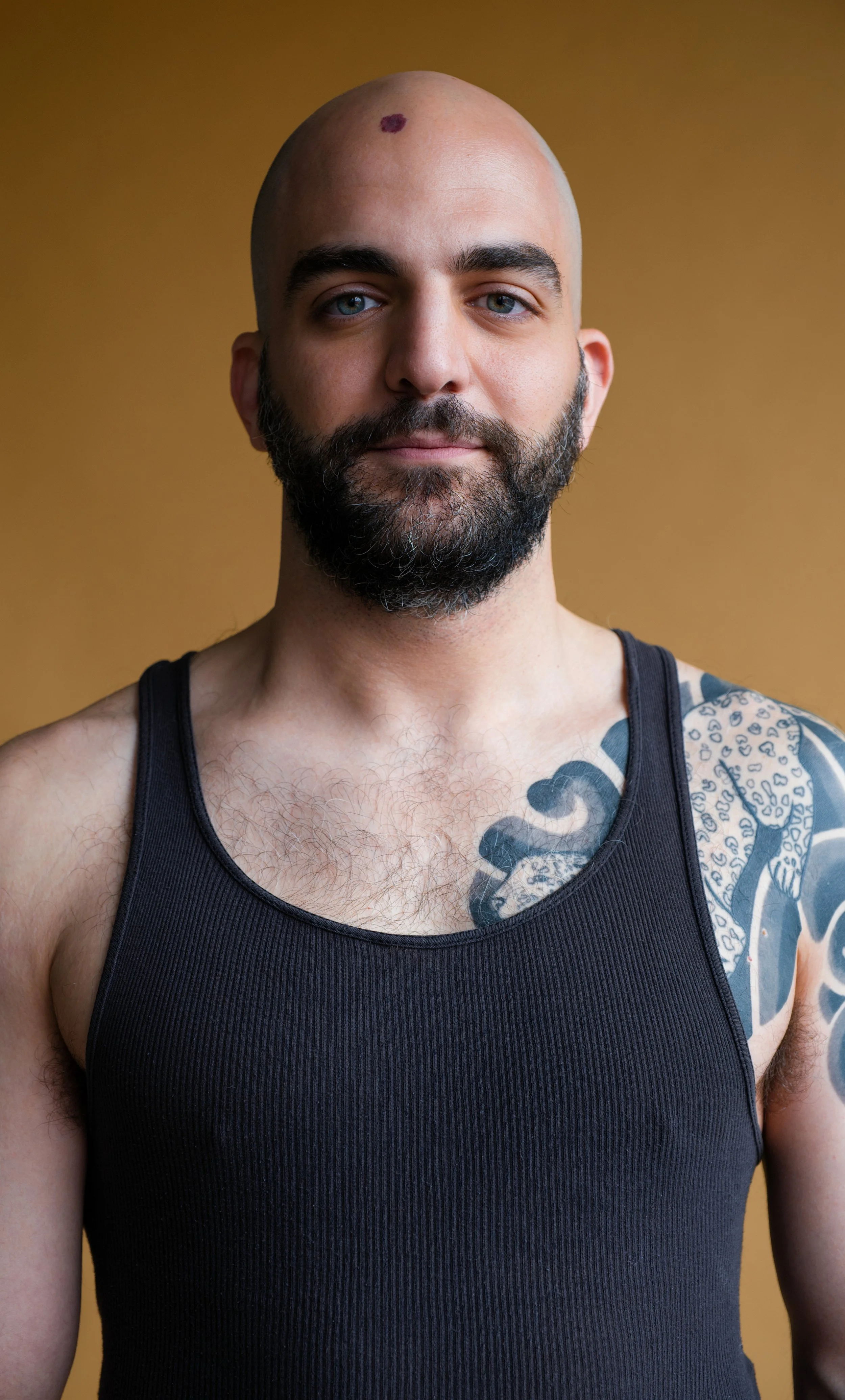 A bald man with a beard, wearing a black tank top and displaying tattoos on his shoulder and chest, poses against a plain brown background.