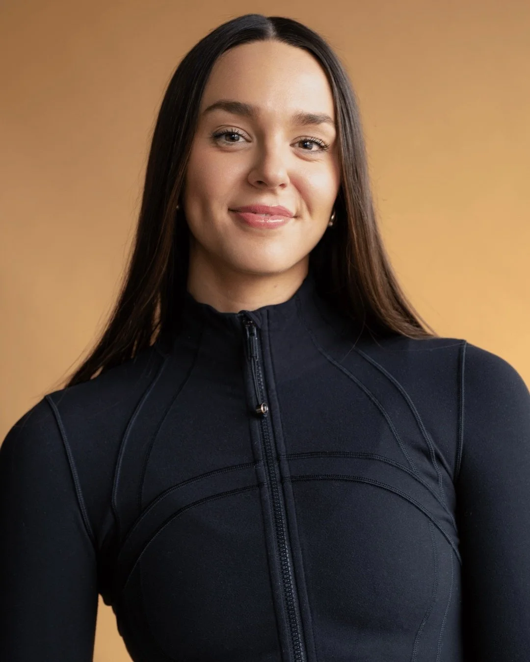 A young woman with long dark hair smiling, wearing a black zip-up athletic top, standing against a warm brown background.