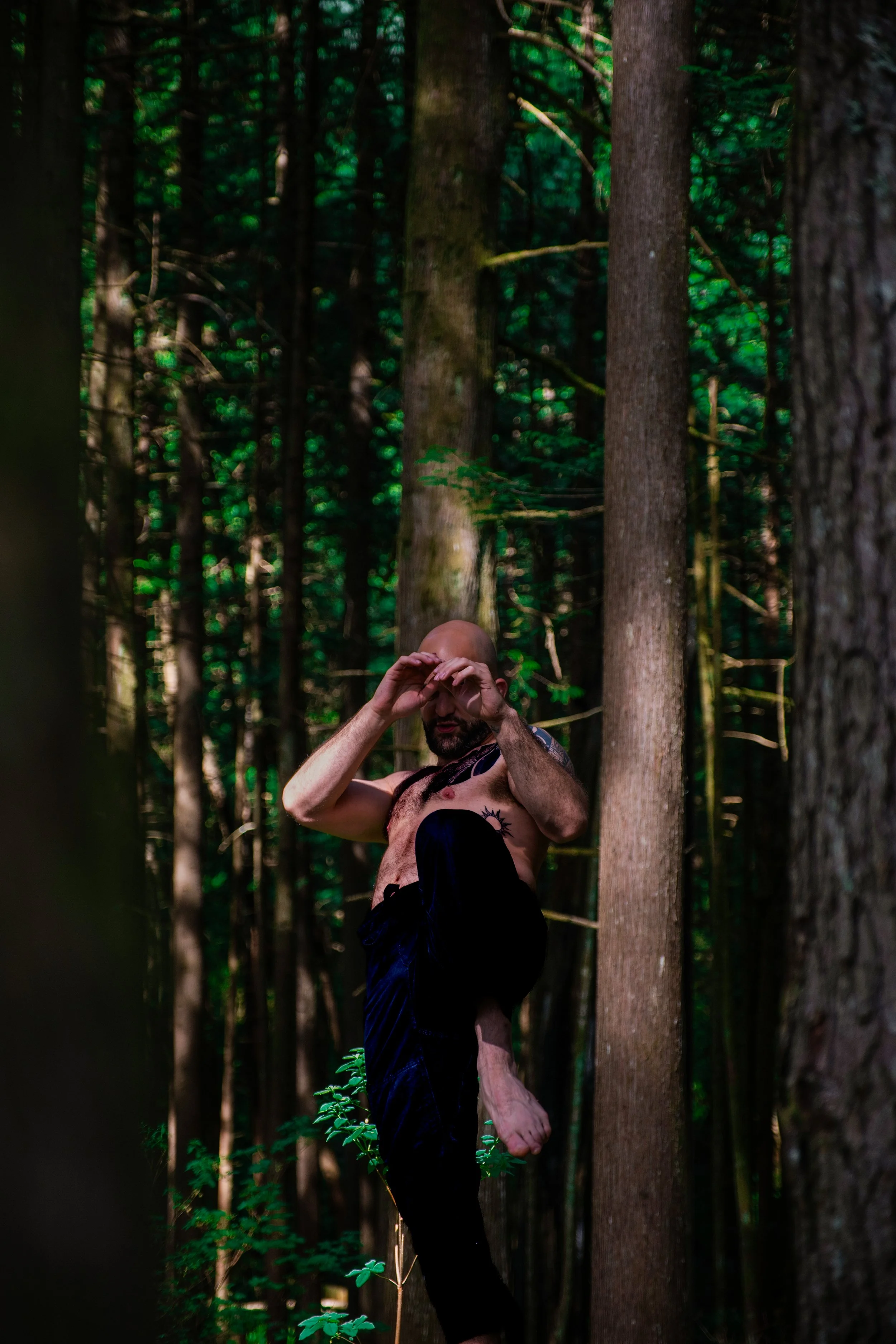 A shirtless man with a tattoo on his chest, standing in a dense forest, lifting his leg high and preforming a martial arts front kick.