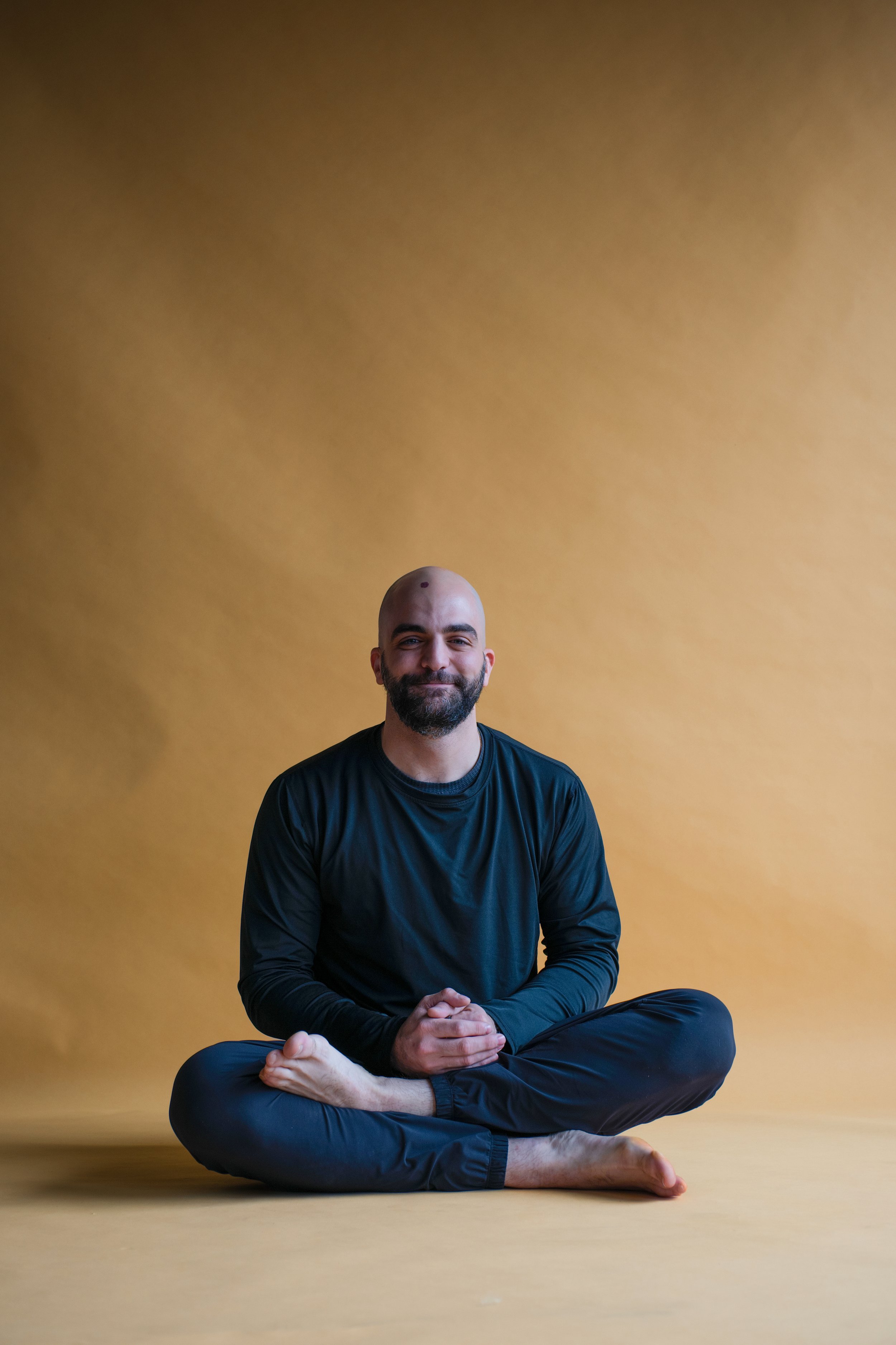 A man sitting cross-legged on the floor against a yellow background, smiling and wearing a black long-sleeve shirt and black pants.