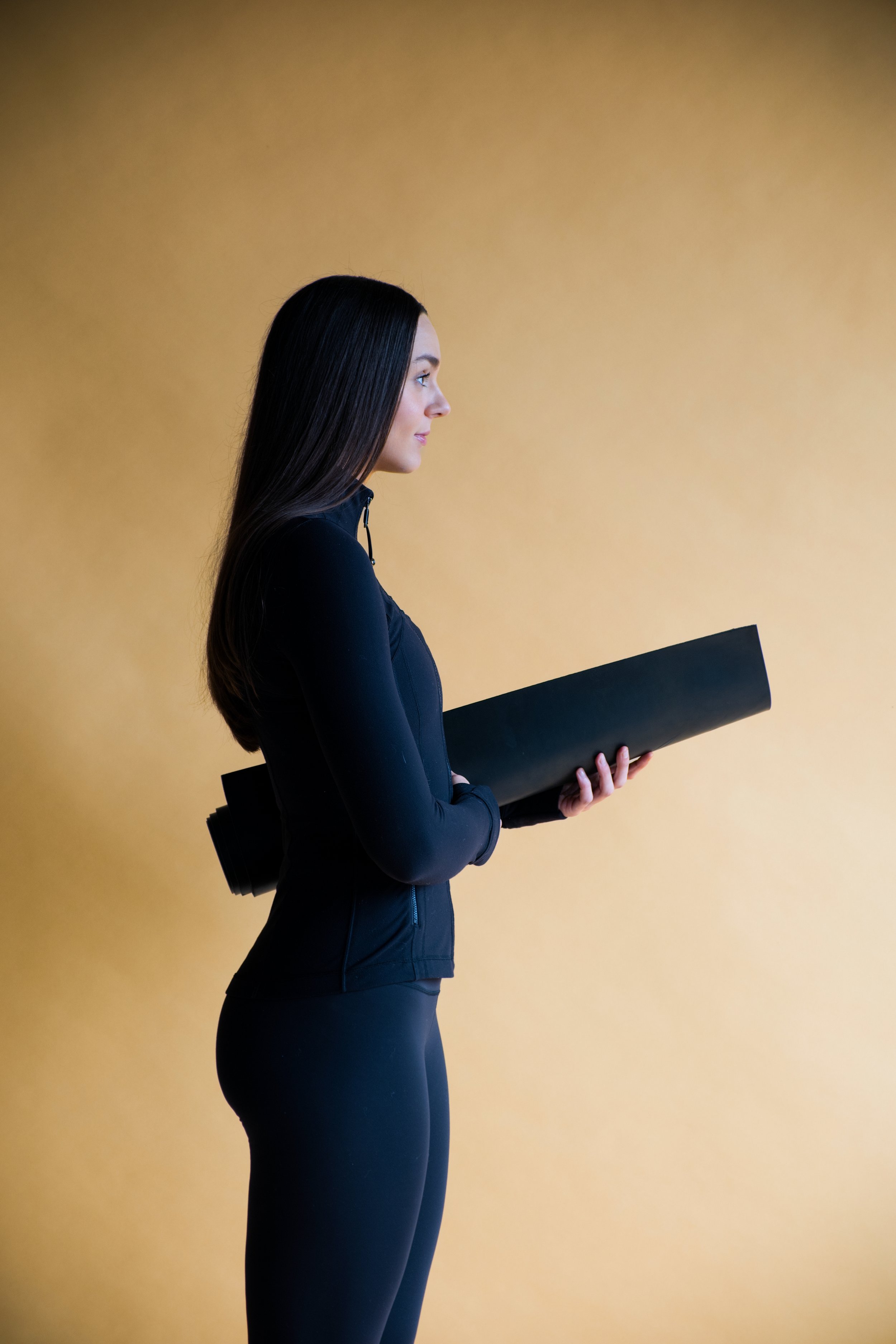 A woman in black athletic clothing holding a rolled-up yoga mat against a yellow background.
