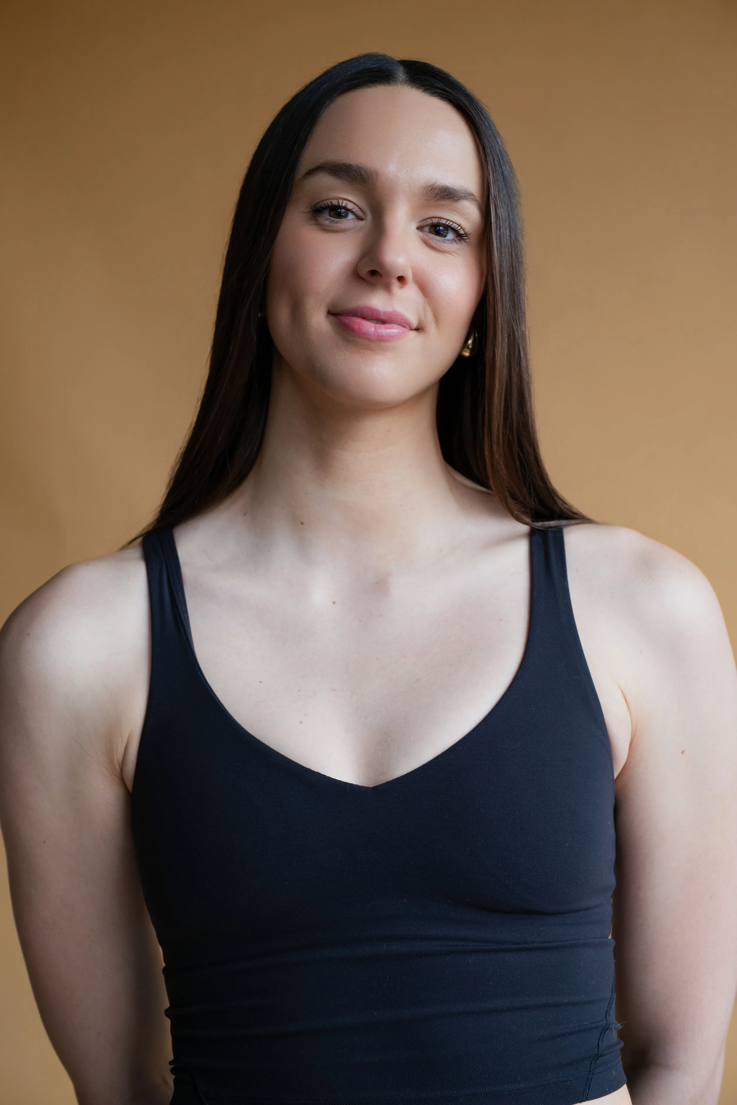 Young woman with long dark hair wearing a black tank top, smiling softly, standing against a plain background.