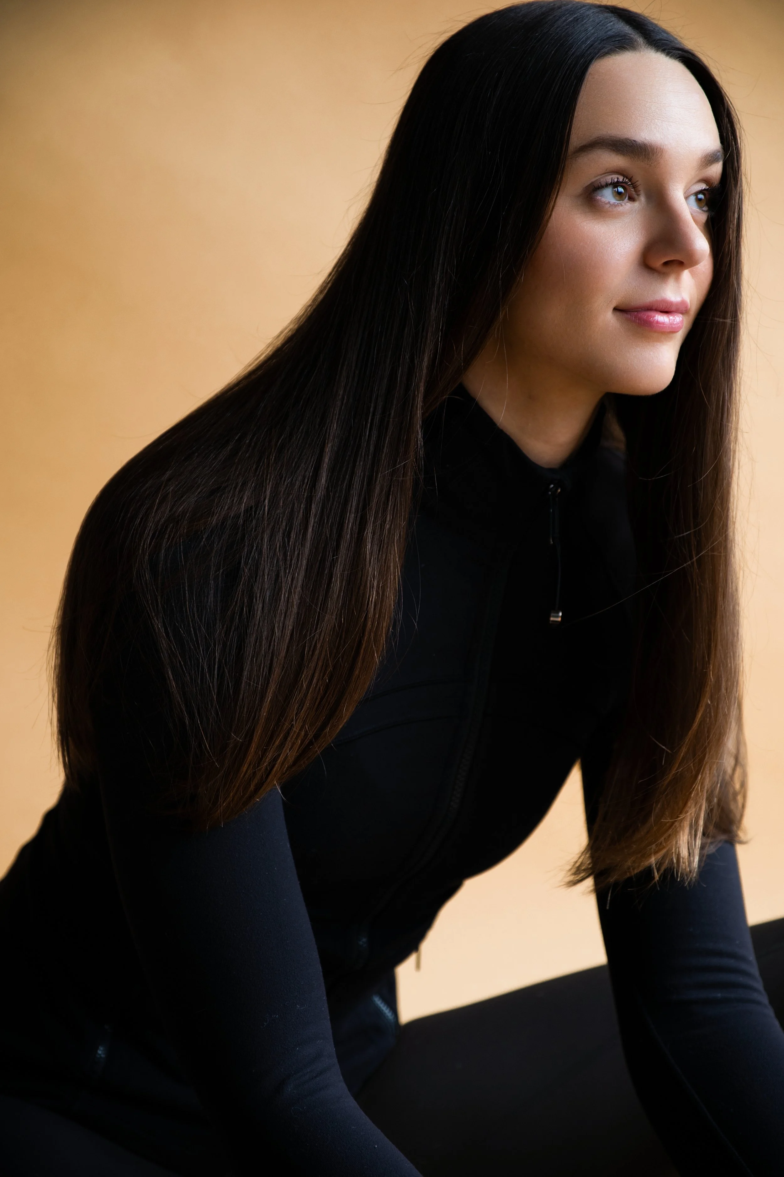 A woman with long brown hair and brown eyes, wearing a black athletic zip-up jacket, looking to the right with a slight smile, against a plain beige background.
