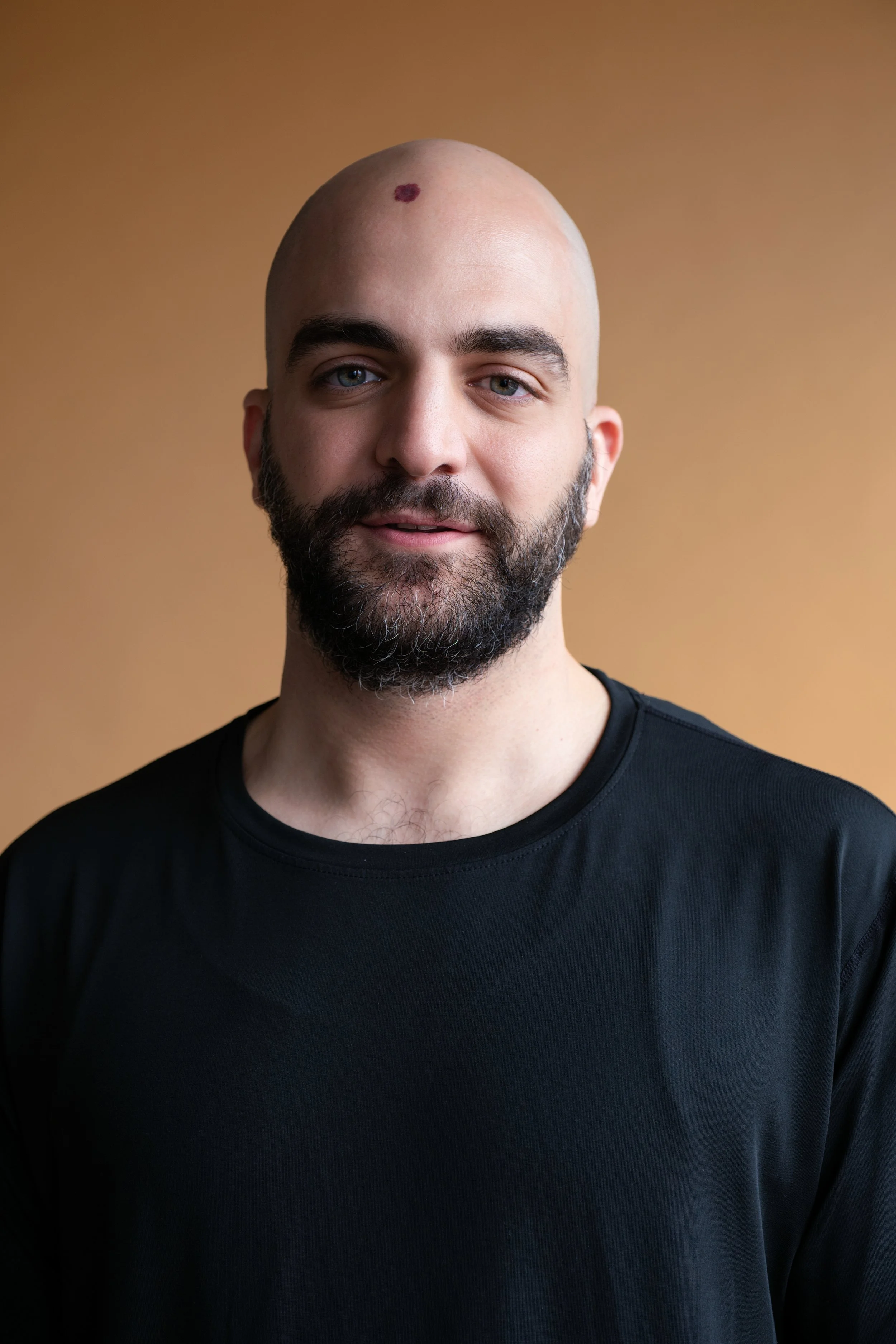 A man with a bald head, blue eyes, and a beard is smiling and wearing a black shirt, standing against a plain brown background.