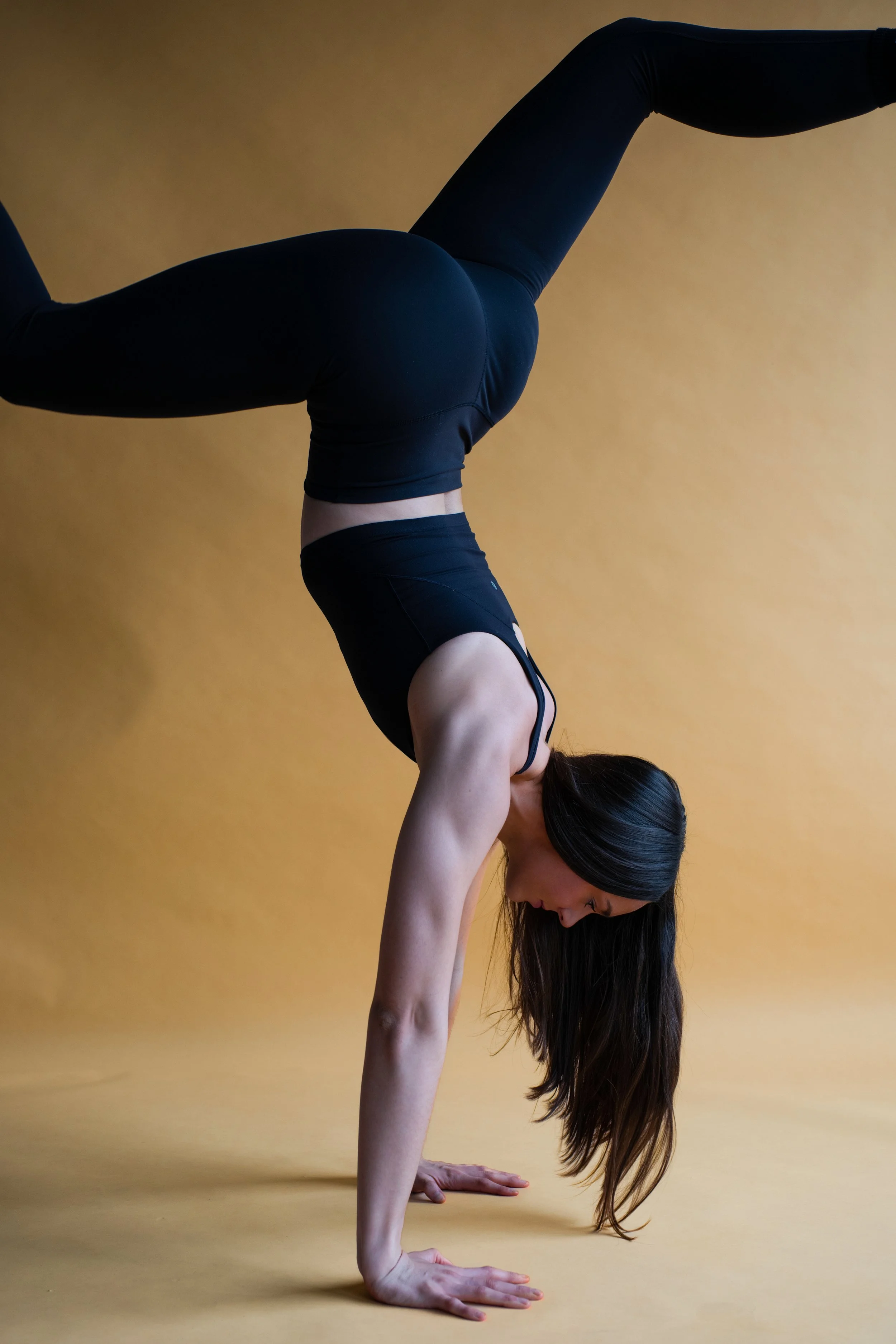 A woman in black activewear performing a handstand against a yellow background.