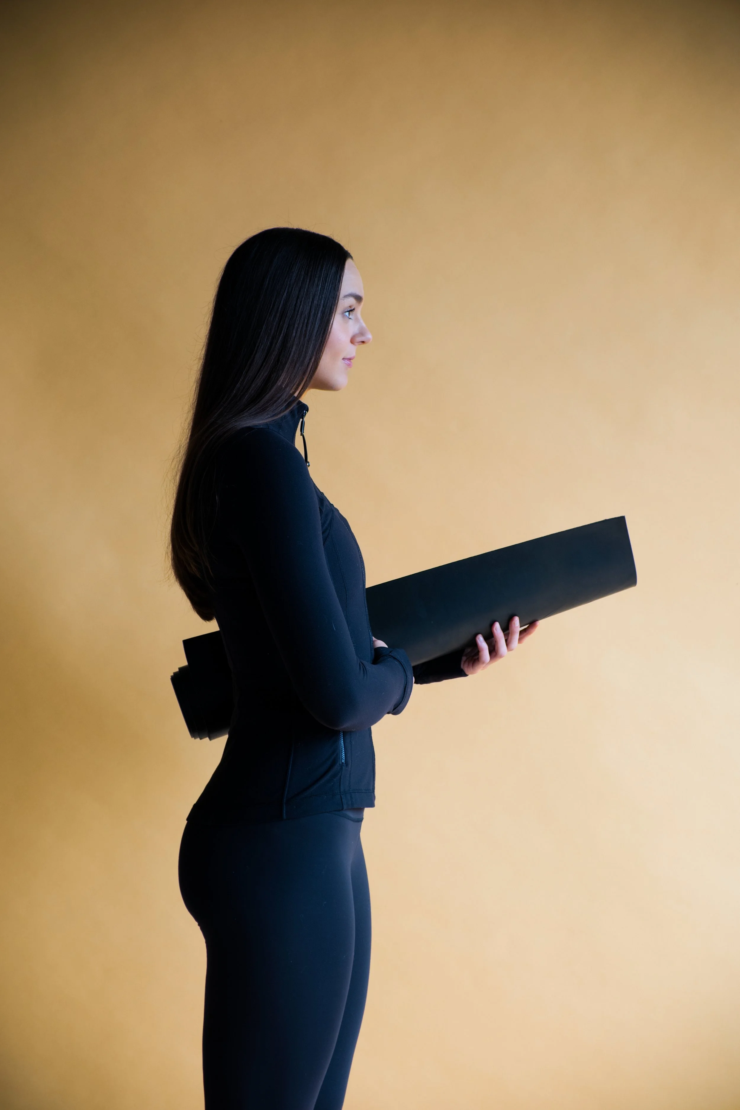 A woman in black athletic clothing holding a yoga mat against a yellow background.