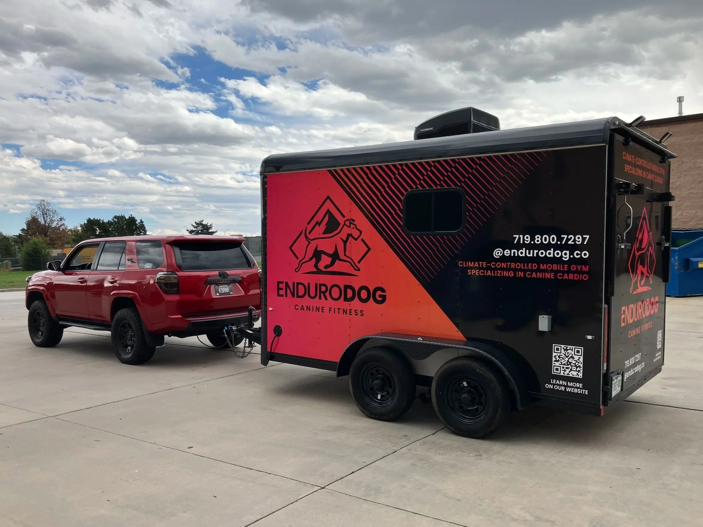 A red SUV connected to a black and red mobile gym trailer with 'EnduroDog' branding, parked on a concrete lot under a partly cloudy sky.