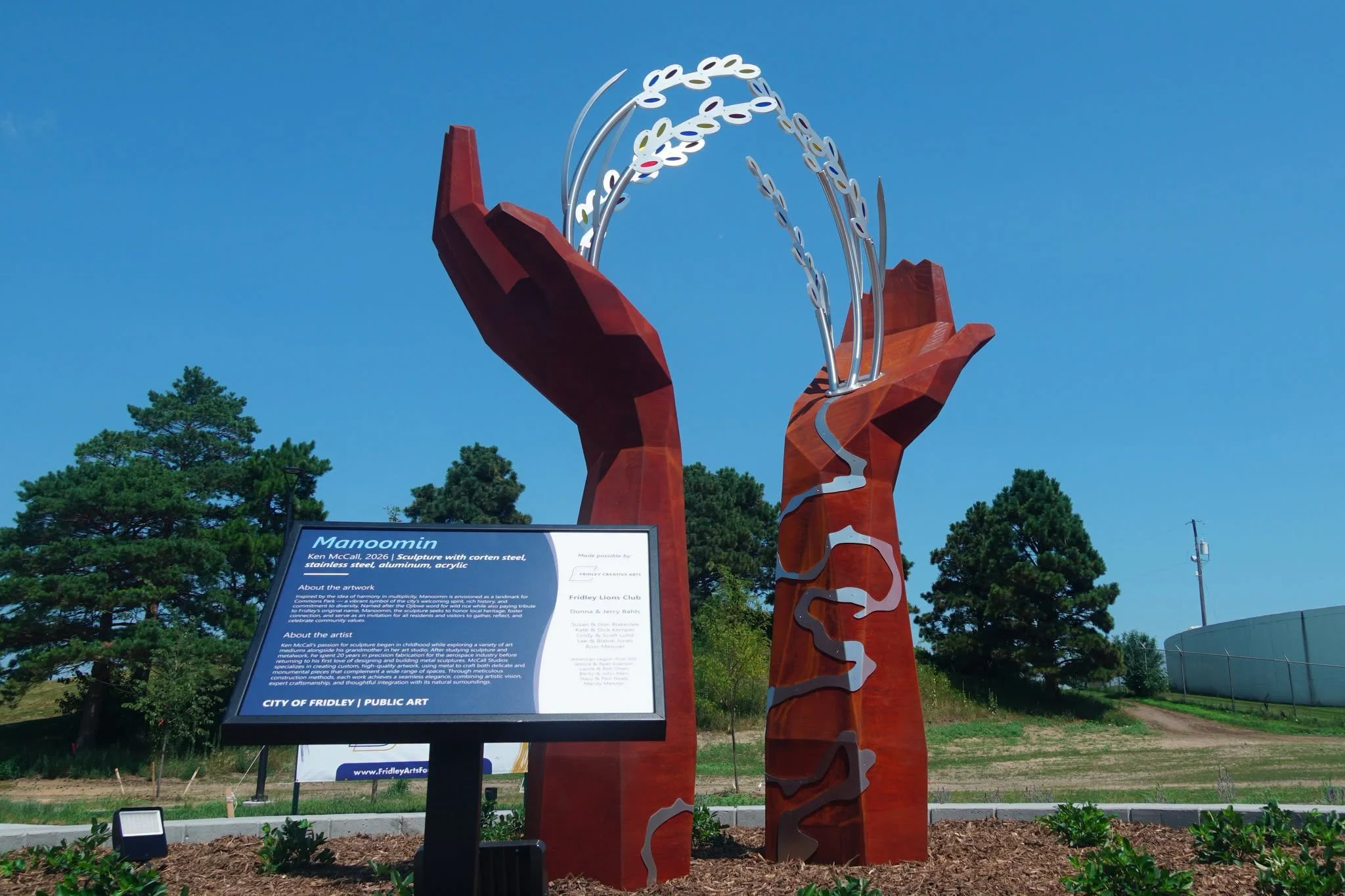 Manoomin public art sculpture at Commons Park in Fridley, featuring two large corten steel hands holding arched wild rice stalks, with an interpretive sign in the foreground.