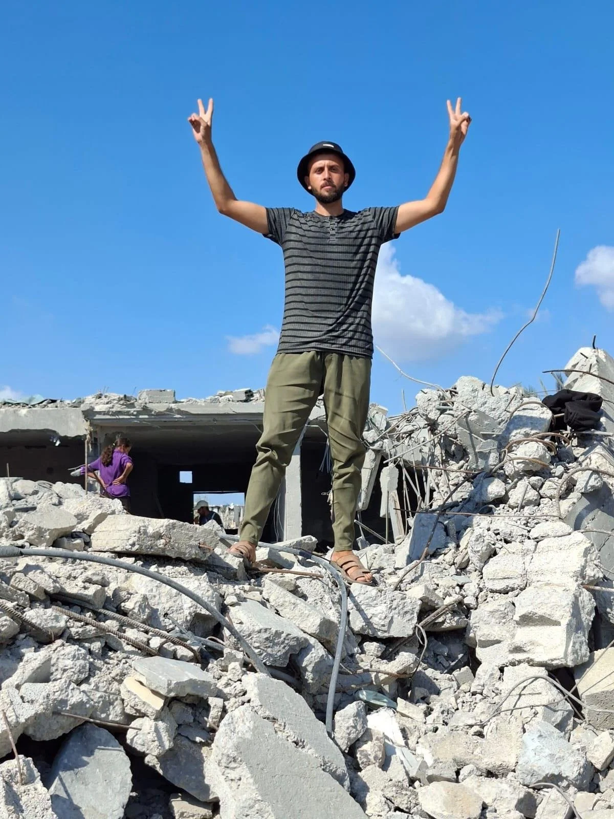 A photo of Tamer standing on top of rubbles, both of his hands raised making "V" signs.