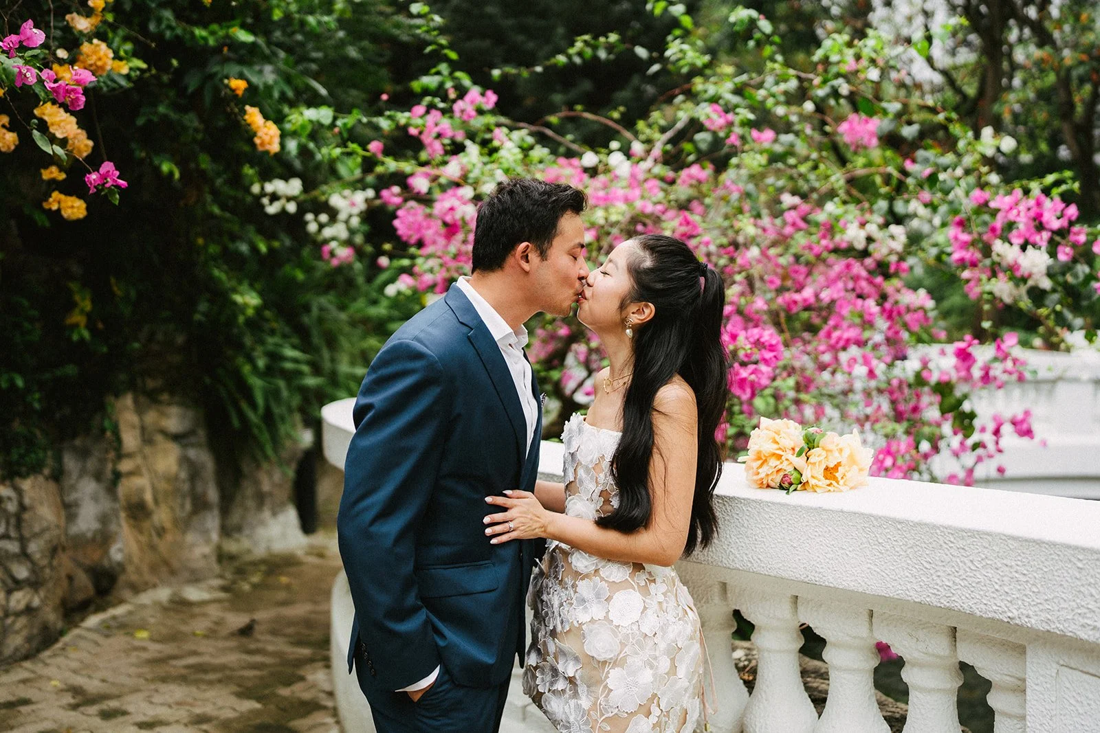 A couple in wedding attire sharing a kiss outdoors with pink and white flowering bushes and a white stone wall in the background.